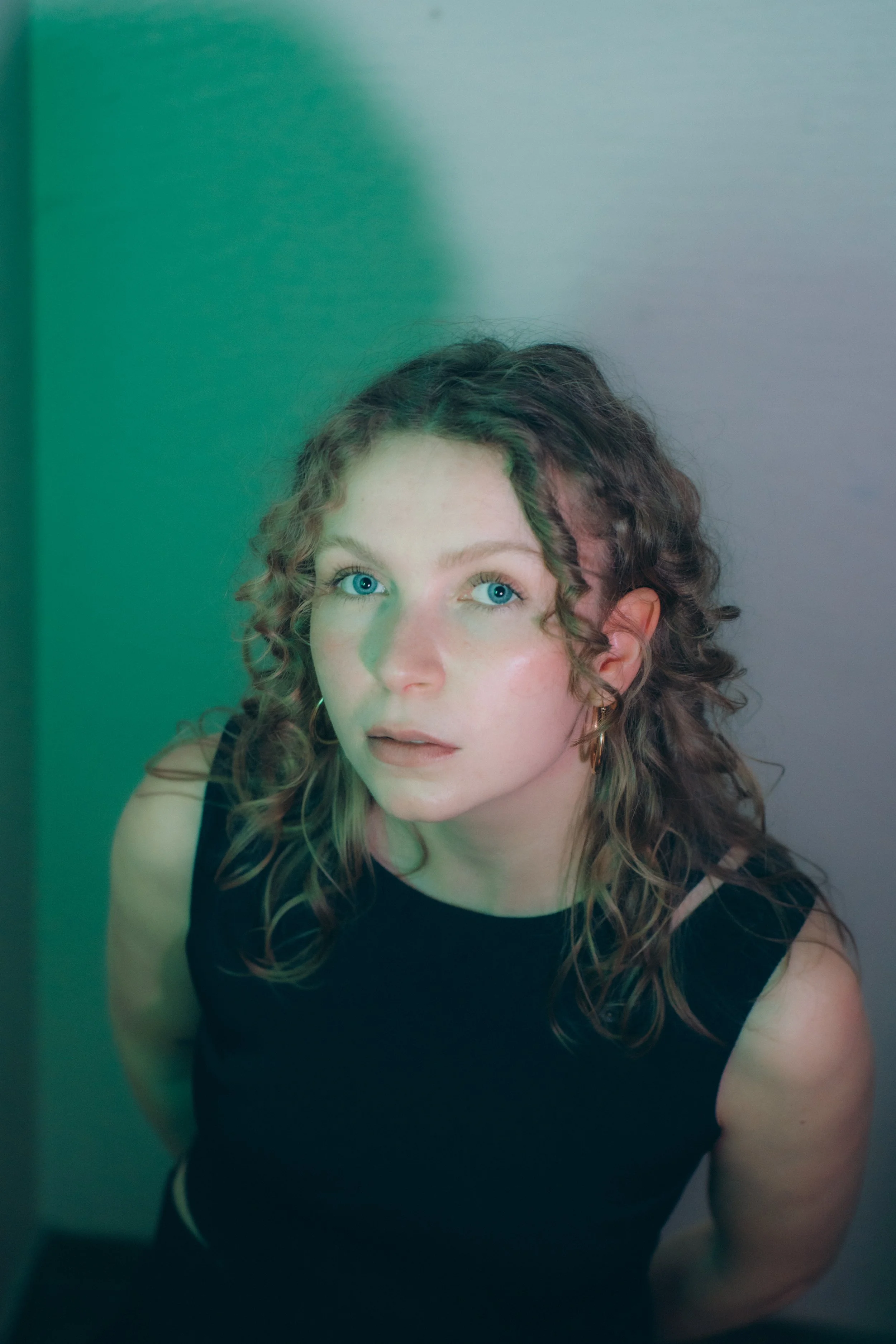 A young woman with curly hair and blue eyes looking directly at the camera. She is wearing a black sleeveless top, gold hoop earrings, and has a neutral facial expression. The background is a plain wall with a subtle gradient.