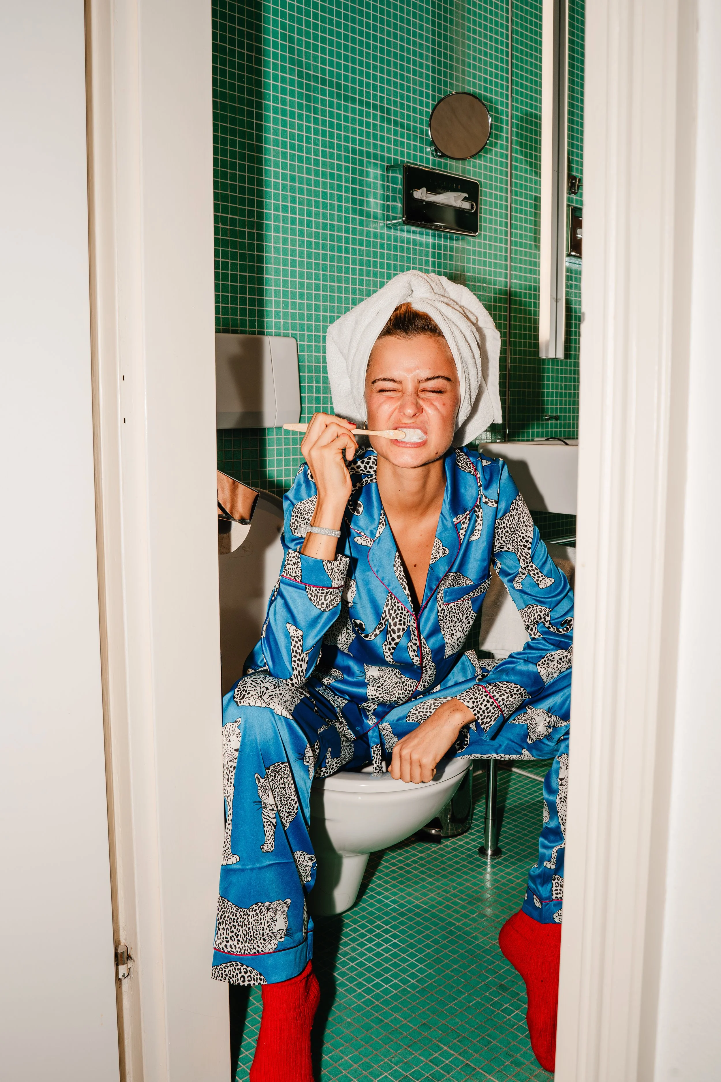 A woman in pajamas with a towel wrapped around her head is sitting on a toilet in a small bathroom. She is holding a toothbrush and appears to be brushing her teeth, with an unhappy expression on her face. The bathroom has green tiles and a mirror ab
