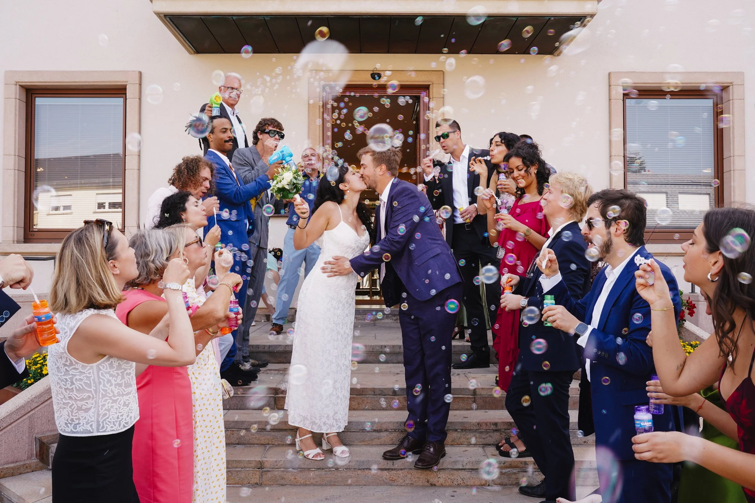 A bride and groom kiss on the steps of a building, surrounded by friends and family celebrating with bubbles and party favors during a wedding.