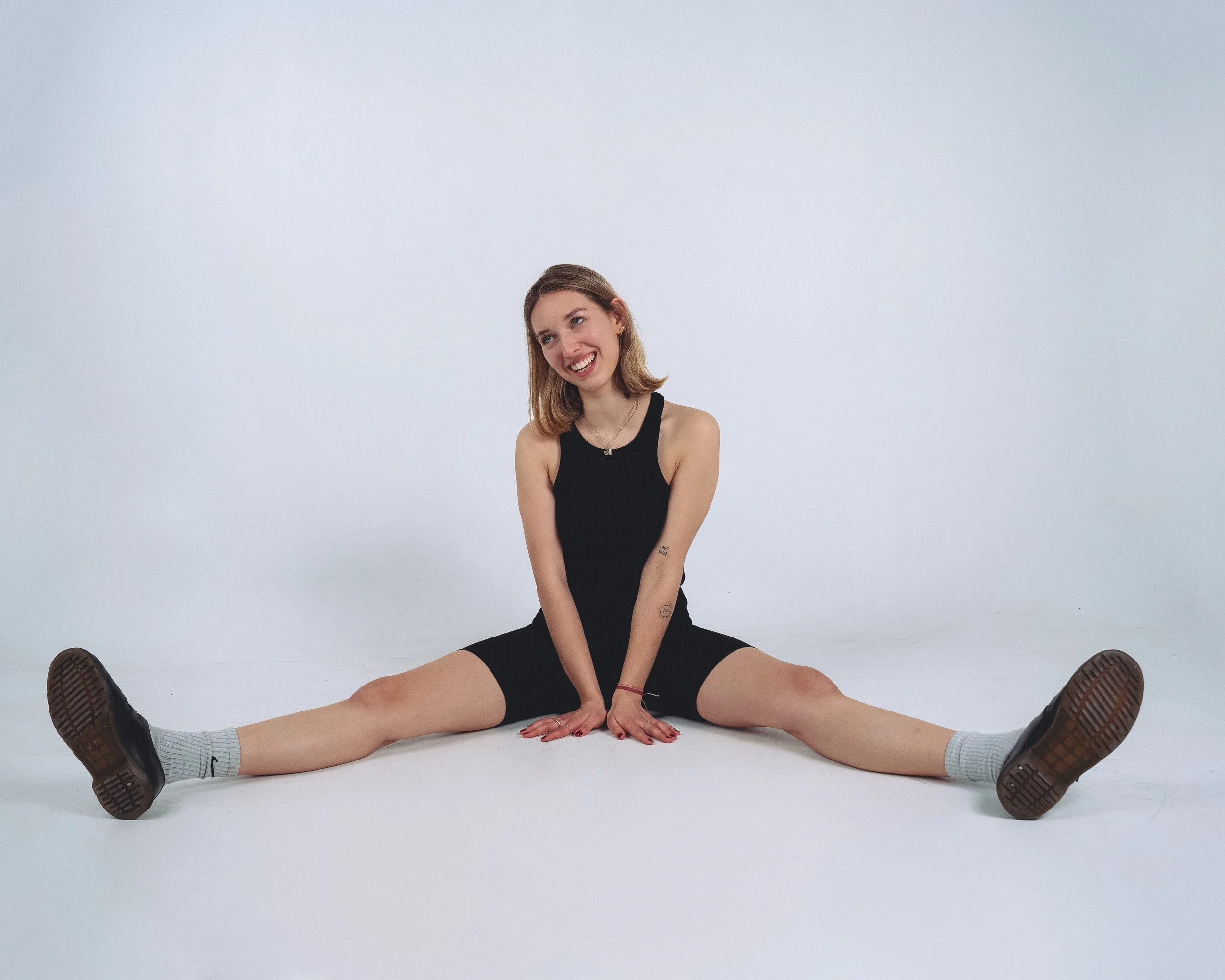 Young woman in black tank top and shorts sitting on the floor in a split pose, smiling and looking to the side with a white background.