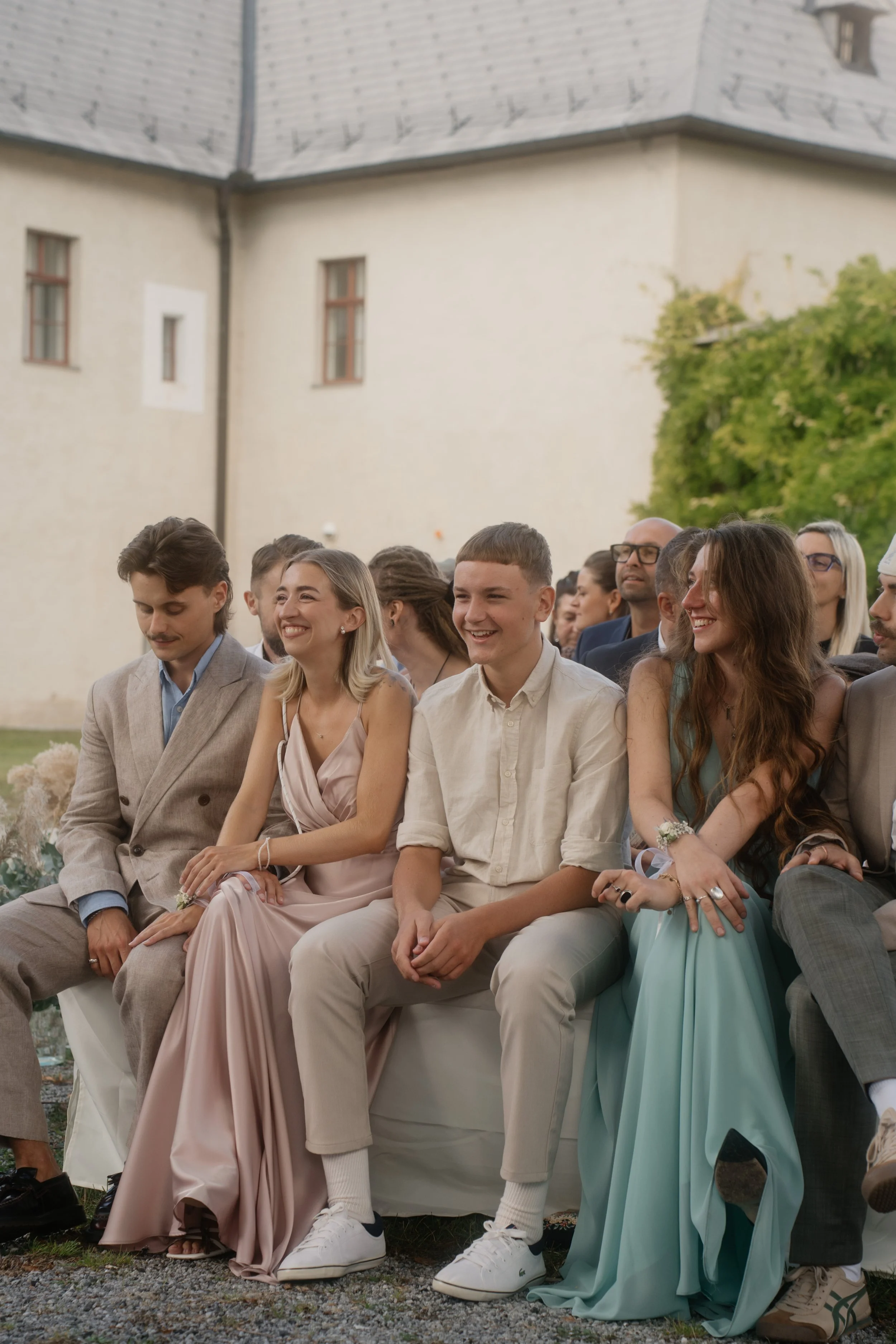 People attending a wedding ceremony seated outdoors in front of a castle-like building. They are smiling and dressed in formal attire, with women in stylish dresses and men in suits.