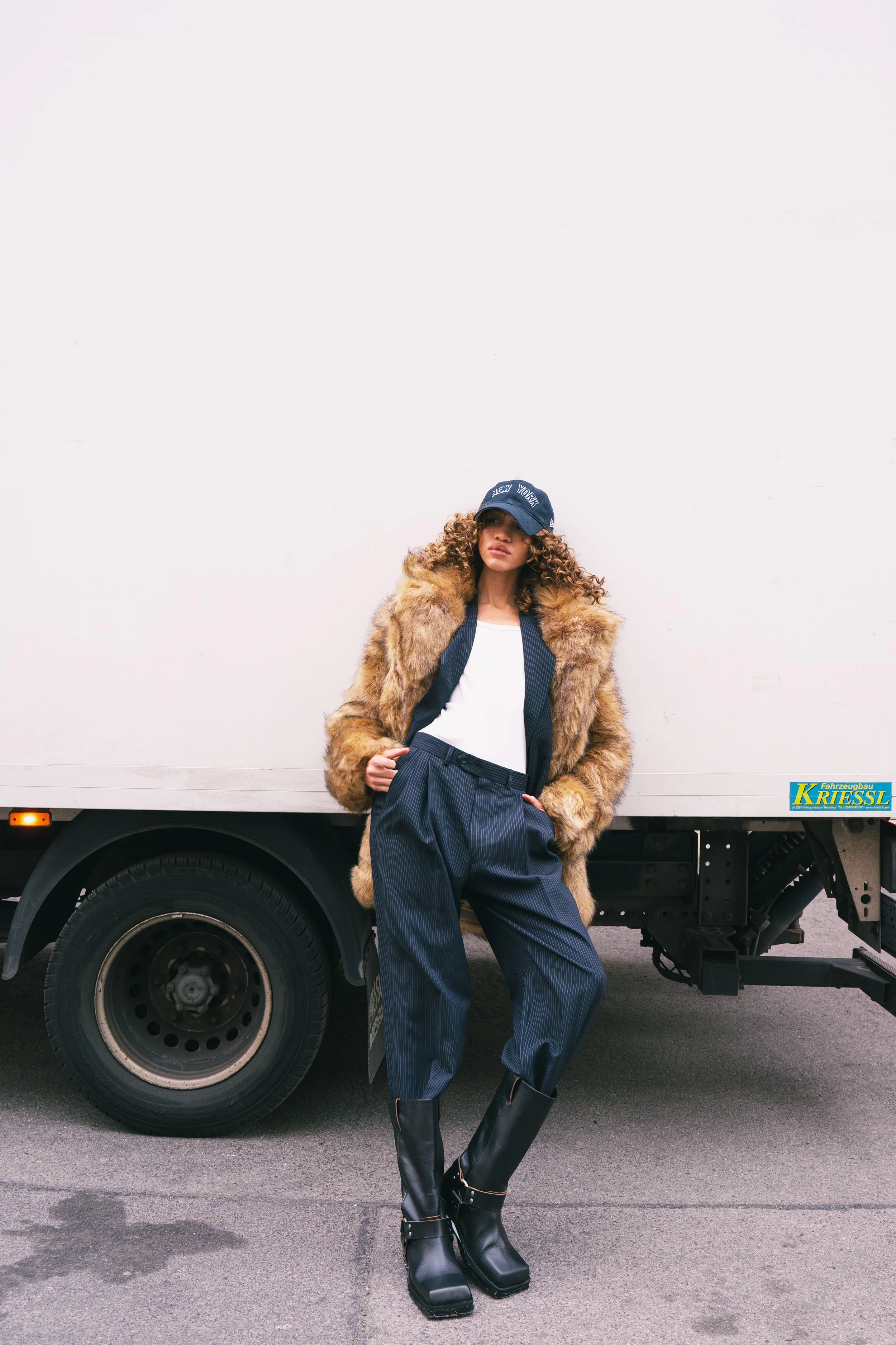 A woman standing in front of a white truck, wearing a fur coat, pinstripe pants, black boots, a white shirt, and a navy cap.