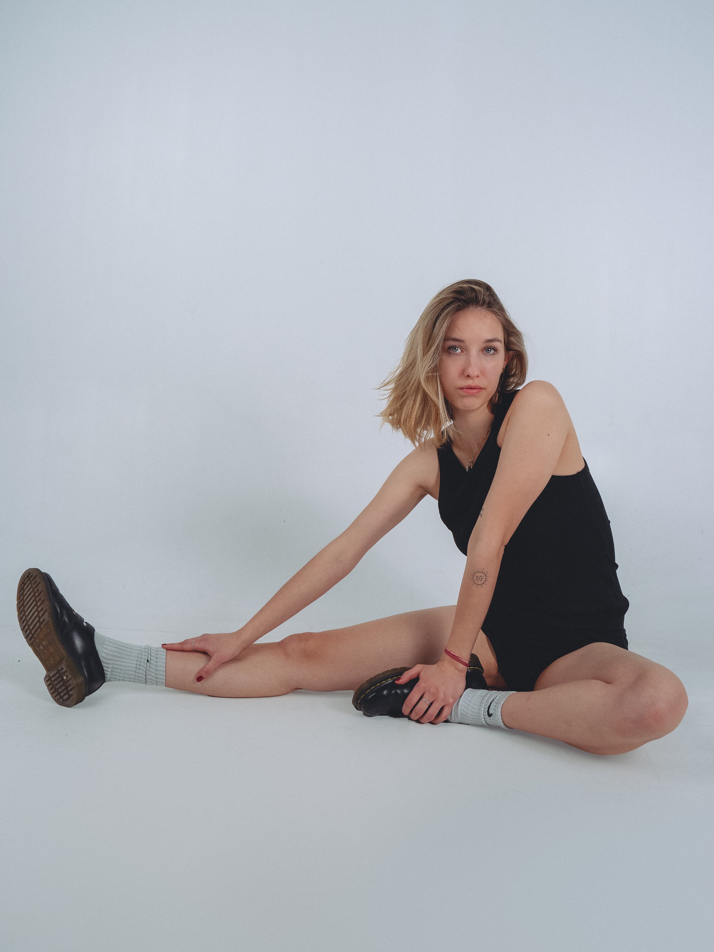 Young woman sitting on the floor in a split position, wearing a black tank top, shorts, grey socks, and black shoes, looking at the camera with a neutral expression.