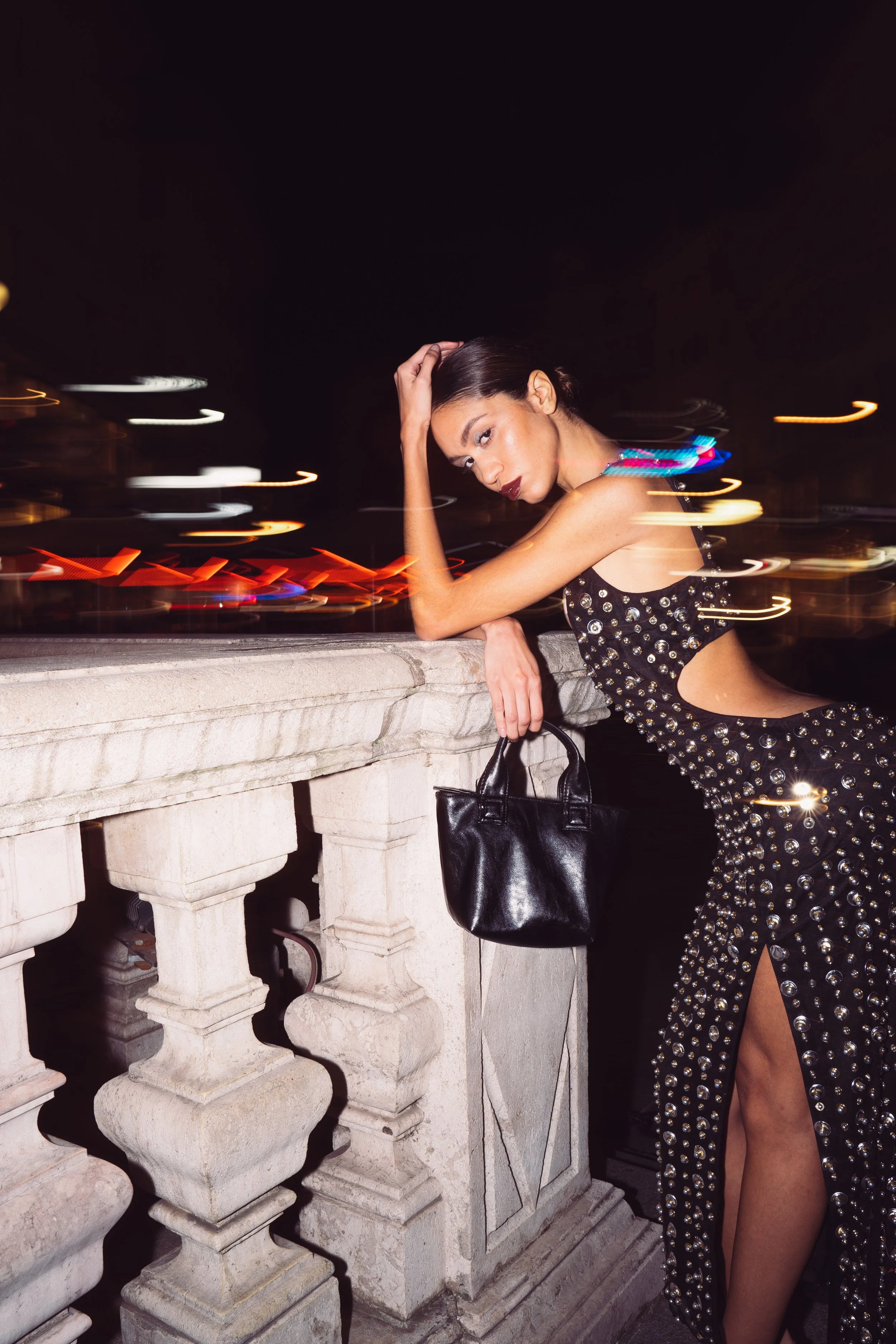 A woman in a black dress with silver embellishments and a high slit leaning on a stone railing at night, holding a black purse, with city lights and motion blur in the background.