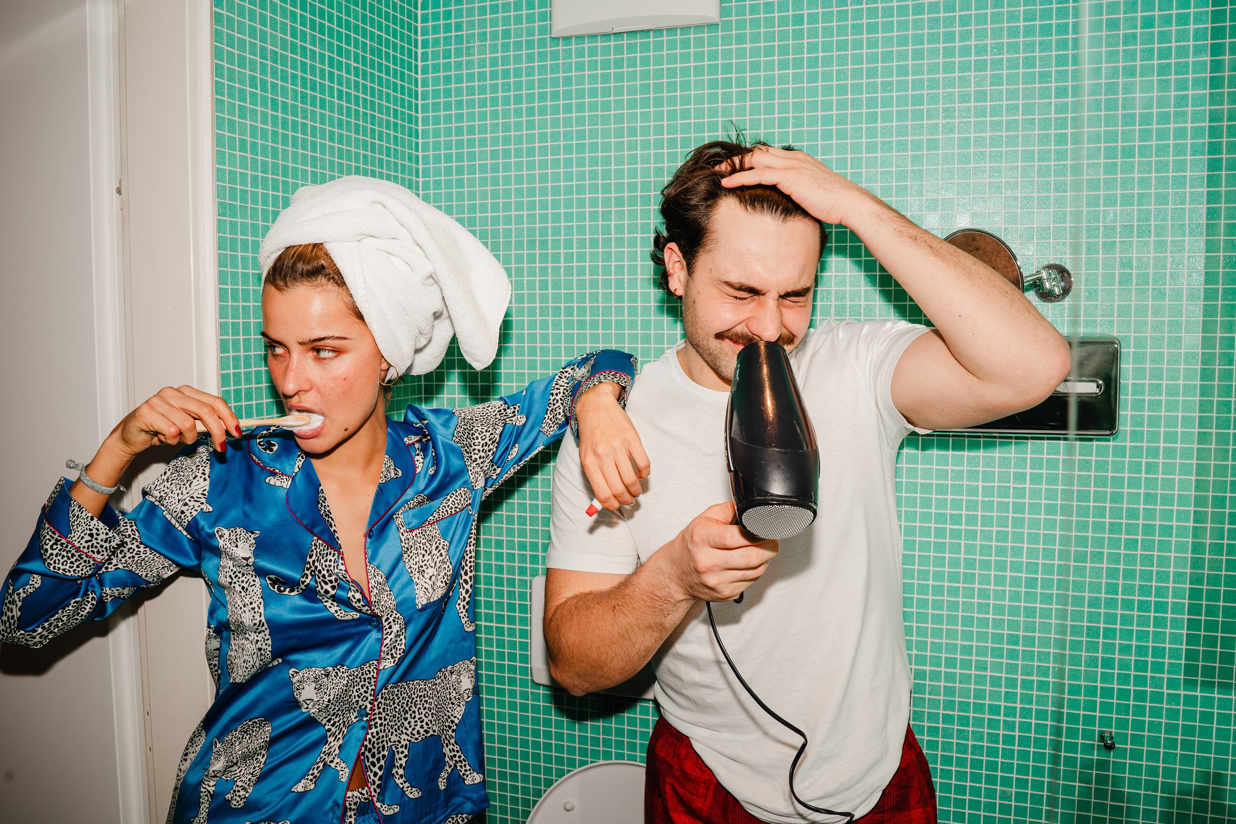 A woman with a towel on her head brushing her teeth while a man dries his hair with a blow dryer in a bathroom with green tiled walls.