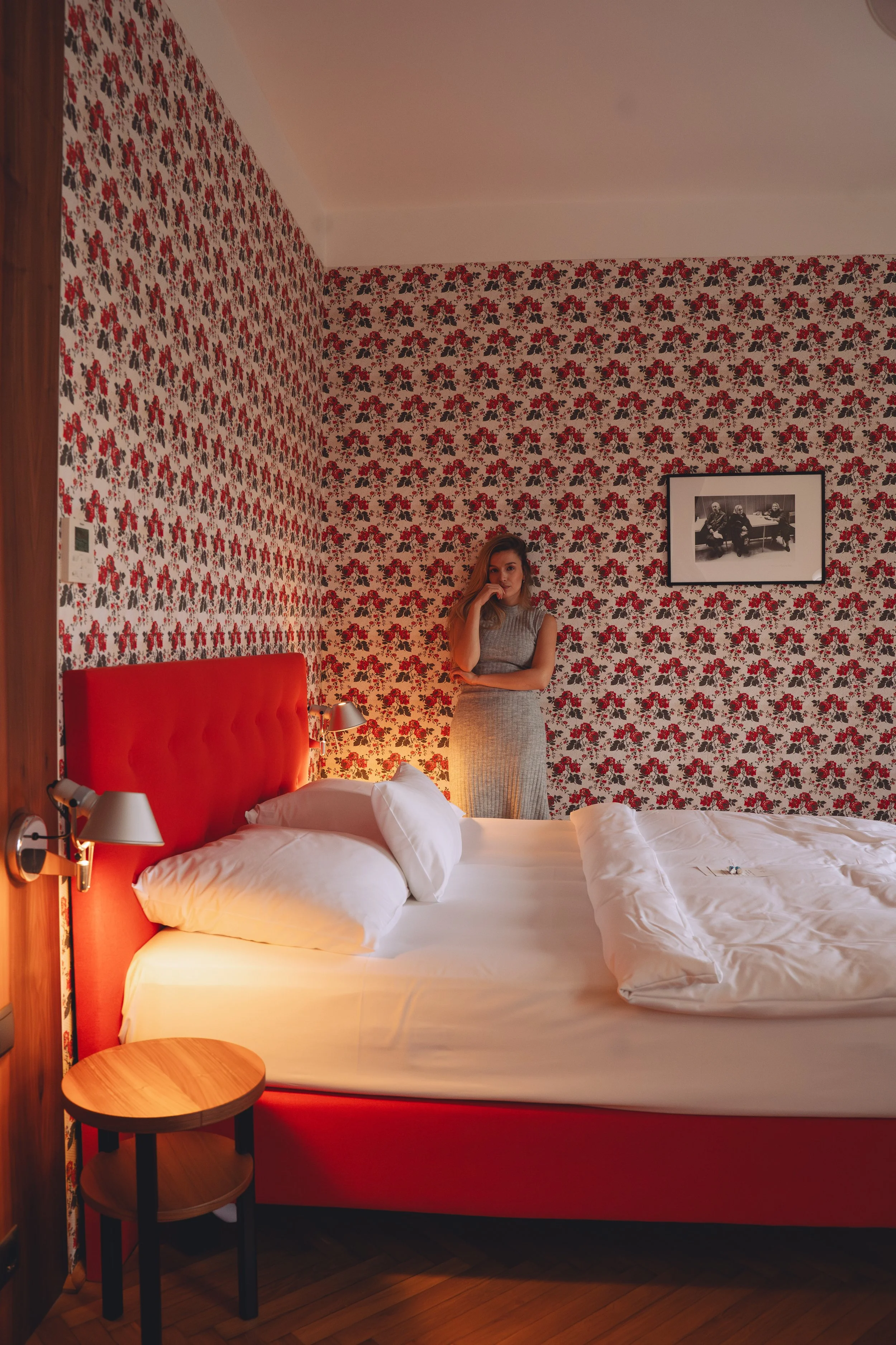 A woman in a patterned grey dress standing beside a bed with a red headboard in a hotel room with floral wallpaper, a small wooden bedside table, and black-and-white picture framed on the wall.