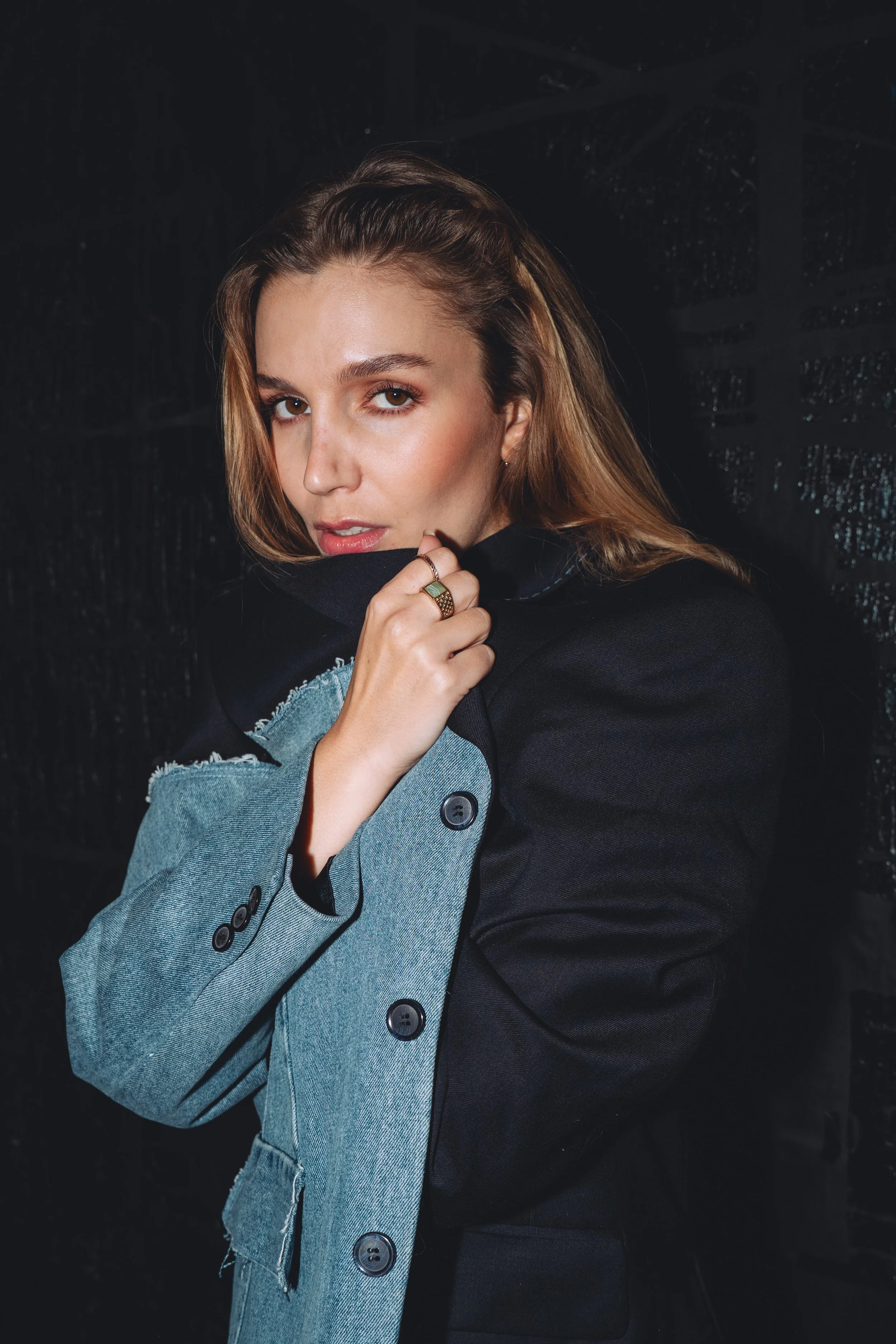 A young woman with long light brown hair, wearing a mixed black and denim jacket, poses against a dark brick wall, holding the collar near her face and looking into the camera.