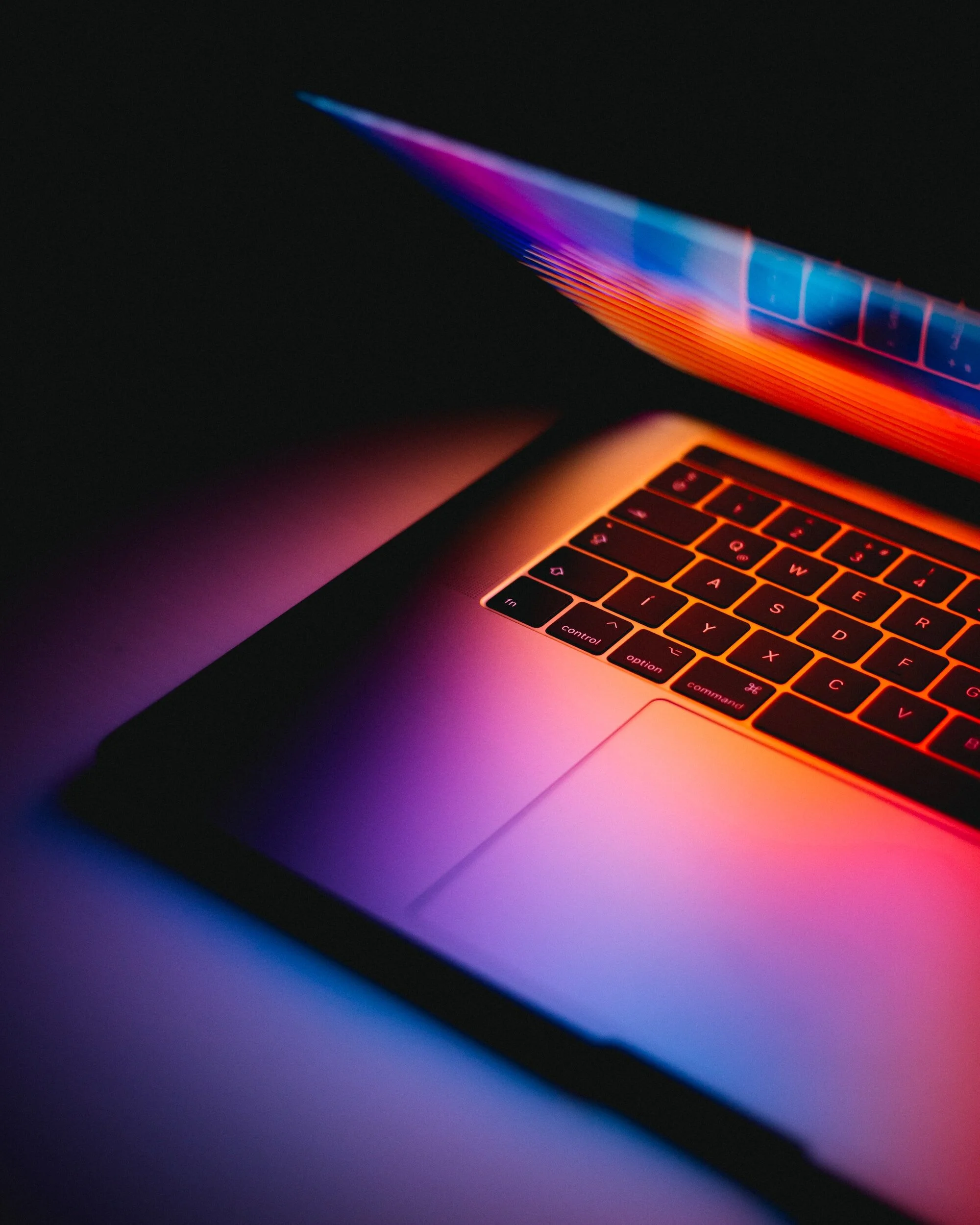 A laptop keyboard and trackpad with colorful light reflections in a dark setting.