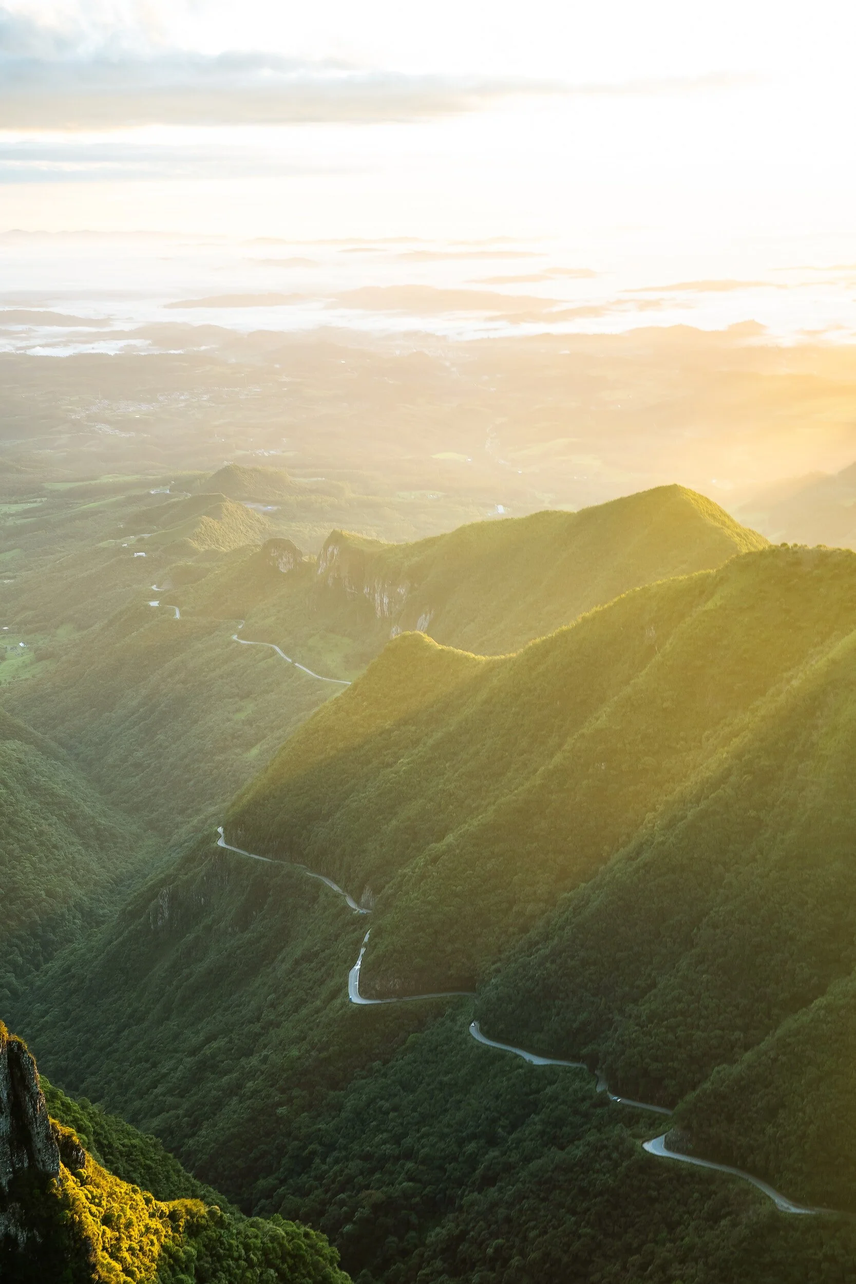 Aerial view of lush green mountain landscape with a winding road, illuminated by warm sunlight.