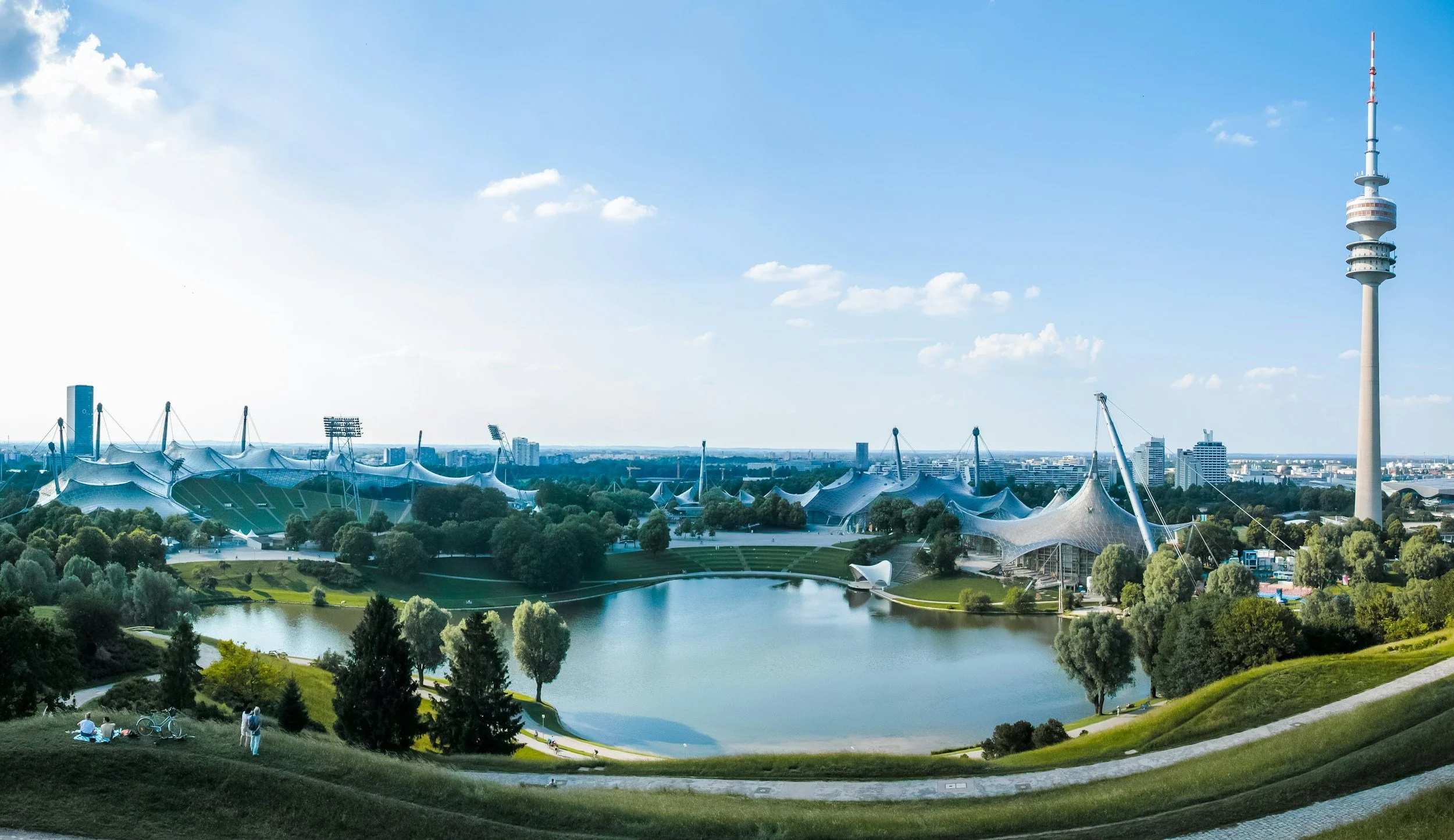 Panoramic view of a park with a lake, futuristic stadium structures, and a tall communications tower on a sunny day.