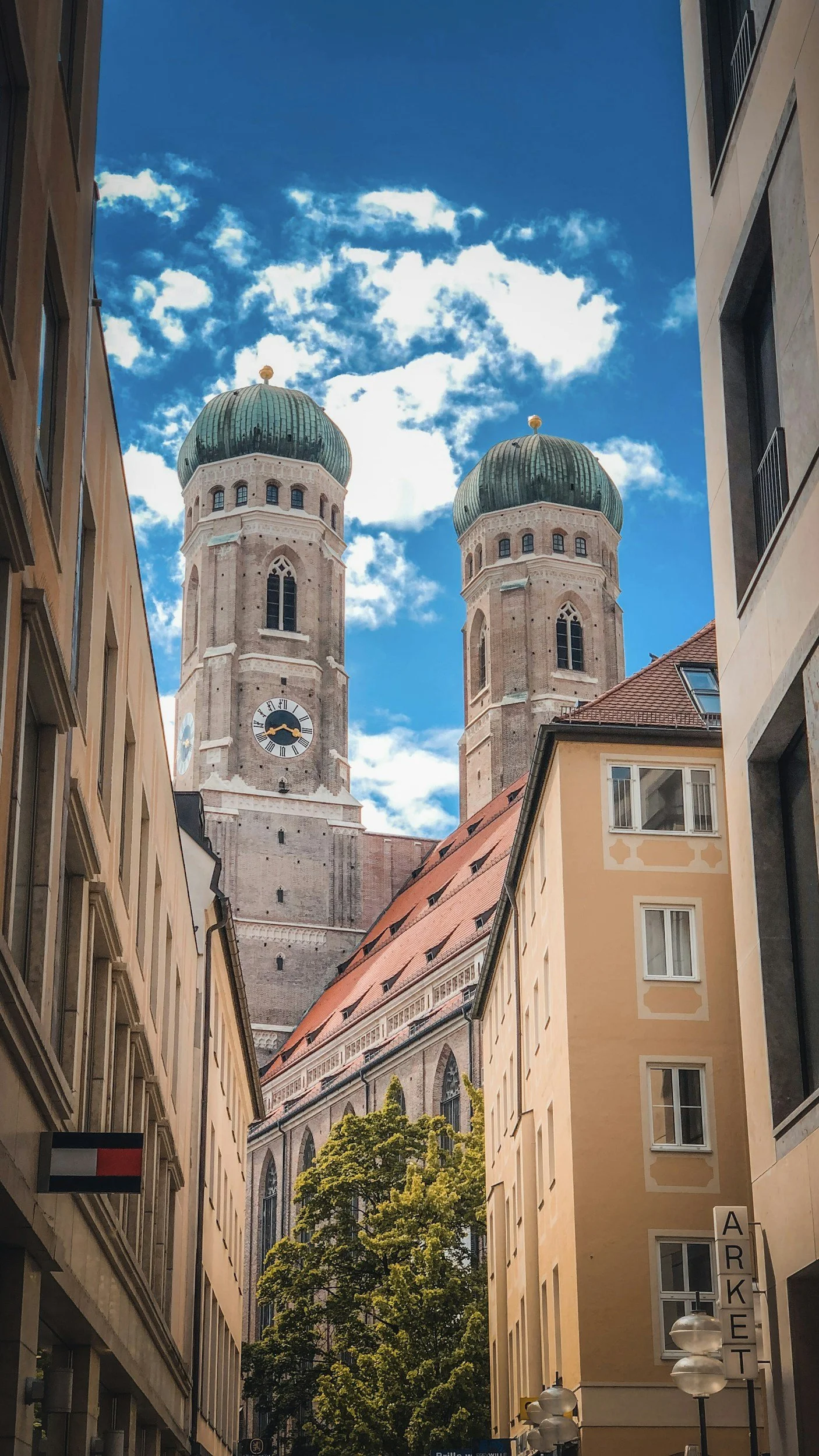 Street view of Frauenkirche cathedral in Munich with its twin green-domed towers, framed by surrounding city buildings under a blue sky.