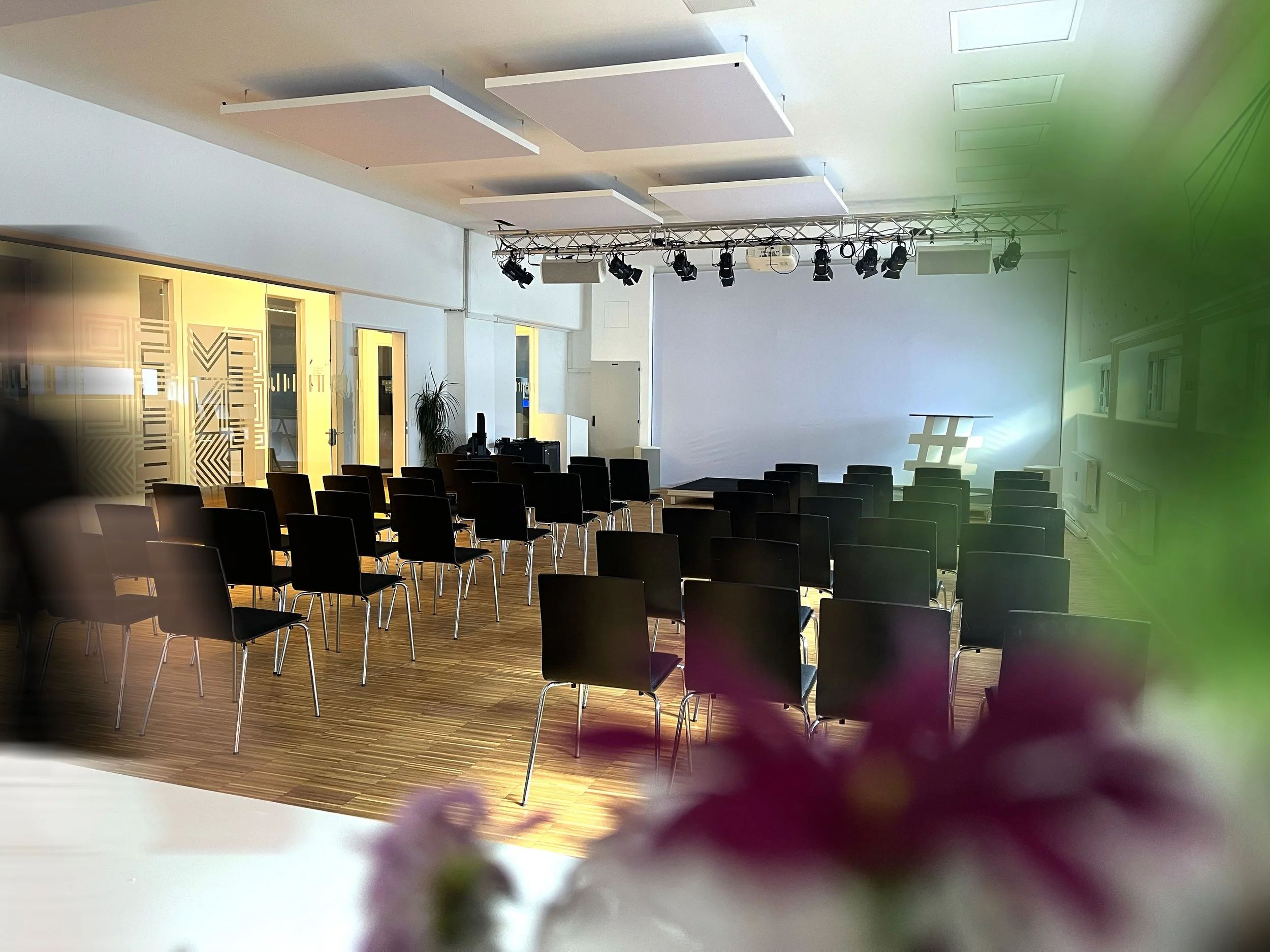 Empty conference room with rows of chairs facing a stage and screen. Overhead lighting fixtures are visible. There are decorative panels on the walls and a blurred plant in the foreground.