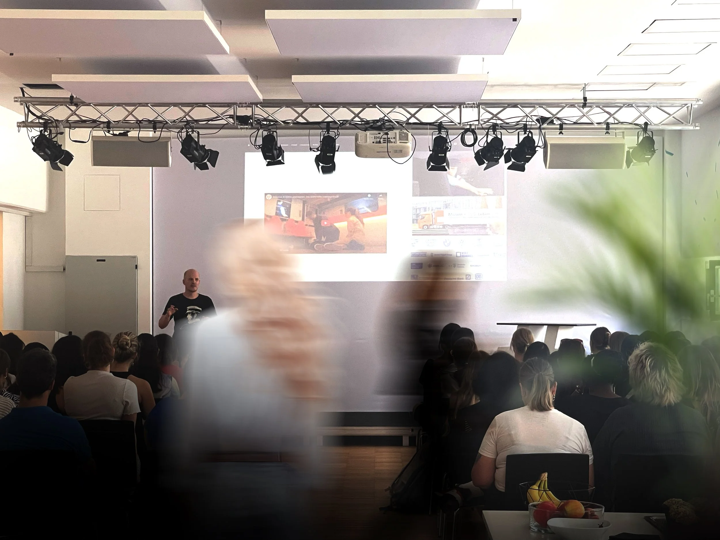 A person giving a presentation to an audience in a dimly lit room. The presenter is standing in front of a projected screen displaying images. The room has professional lighting equipment, and there is a small table with fruit in the foreground. Some people in the audience are slightly blurred, indicating motion.