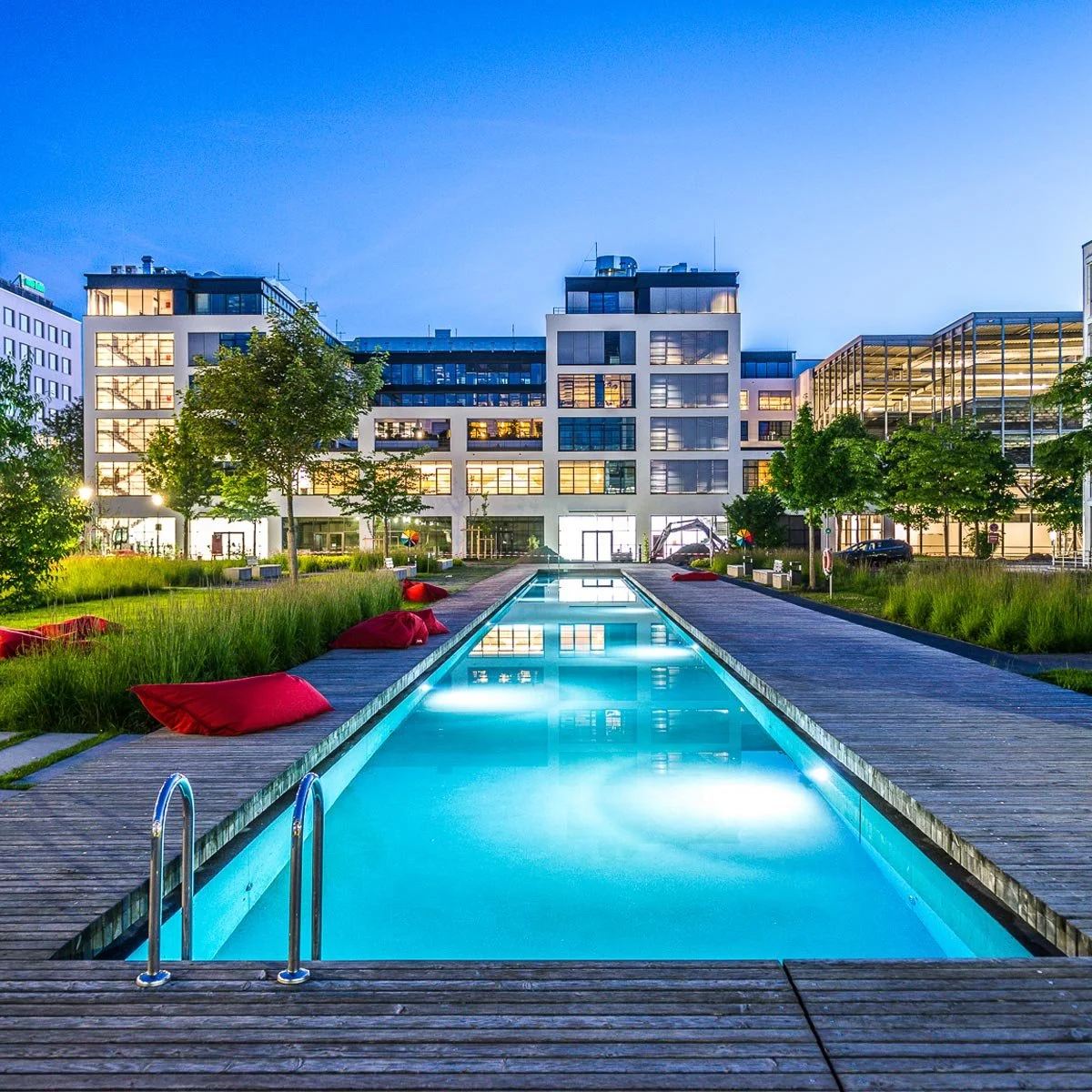 Modern building with illuminated windows behind a long rectangular pool, flanked by wooden decking and grassy areas with red cushions, at twilight.