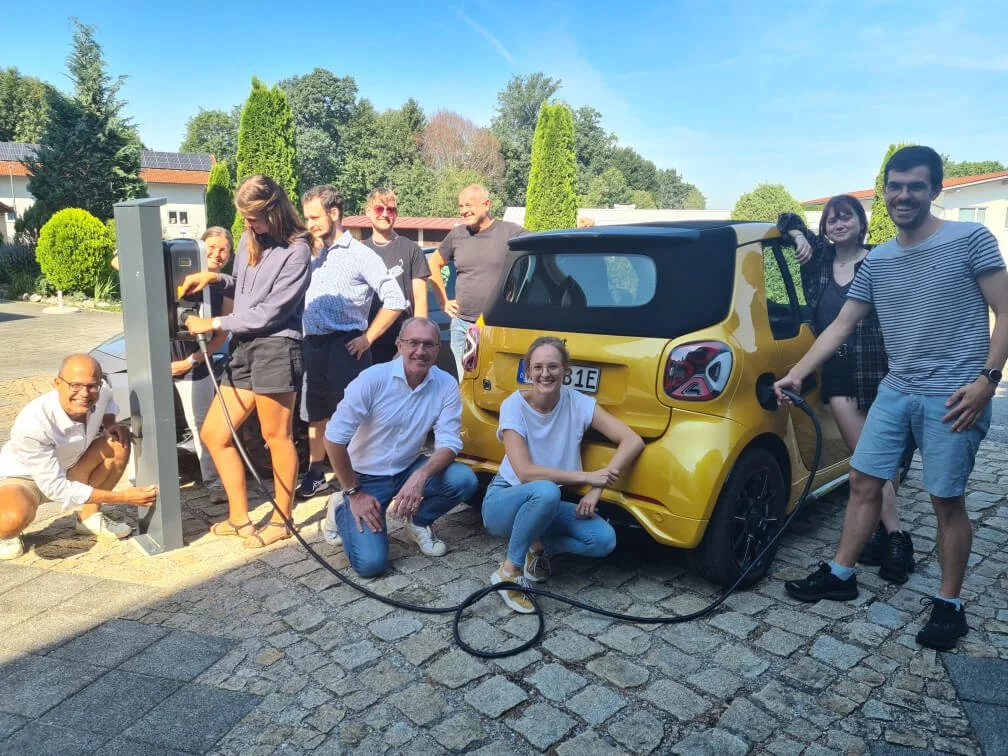Group of people gathered around a yellow electric car, charging it outdoors. The setting is a sunny day with a backdrop of greenery and houses.