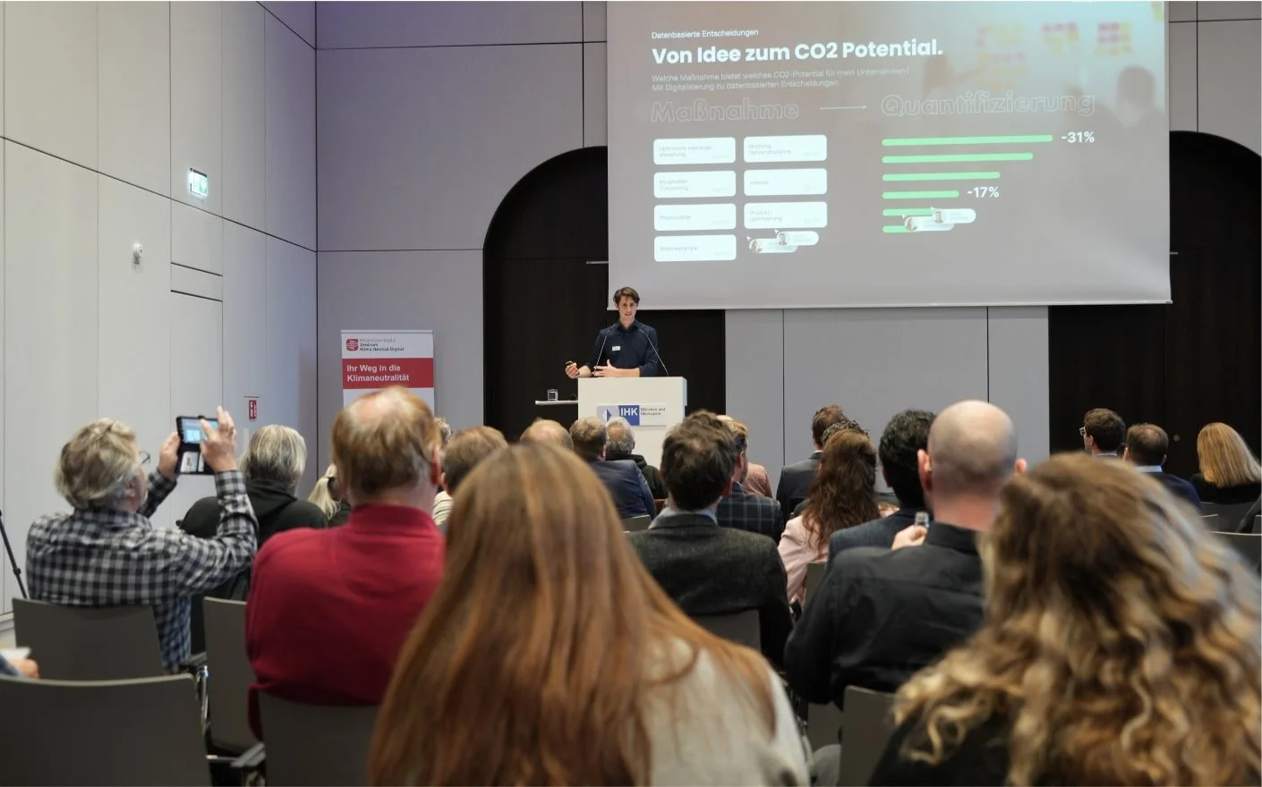 A man giving a presentation about CO2 potential at an event with an audience watching. A projection screen displays information in German with charts and graphs. Attendees sit in rows, some taking notes or photos.