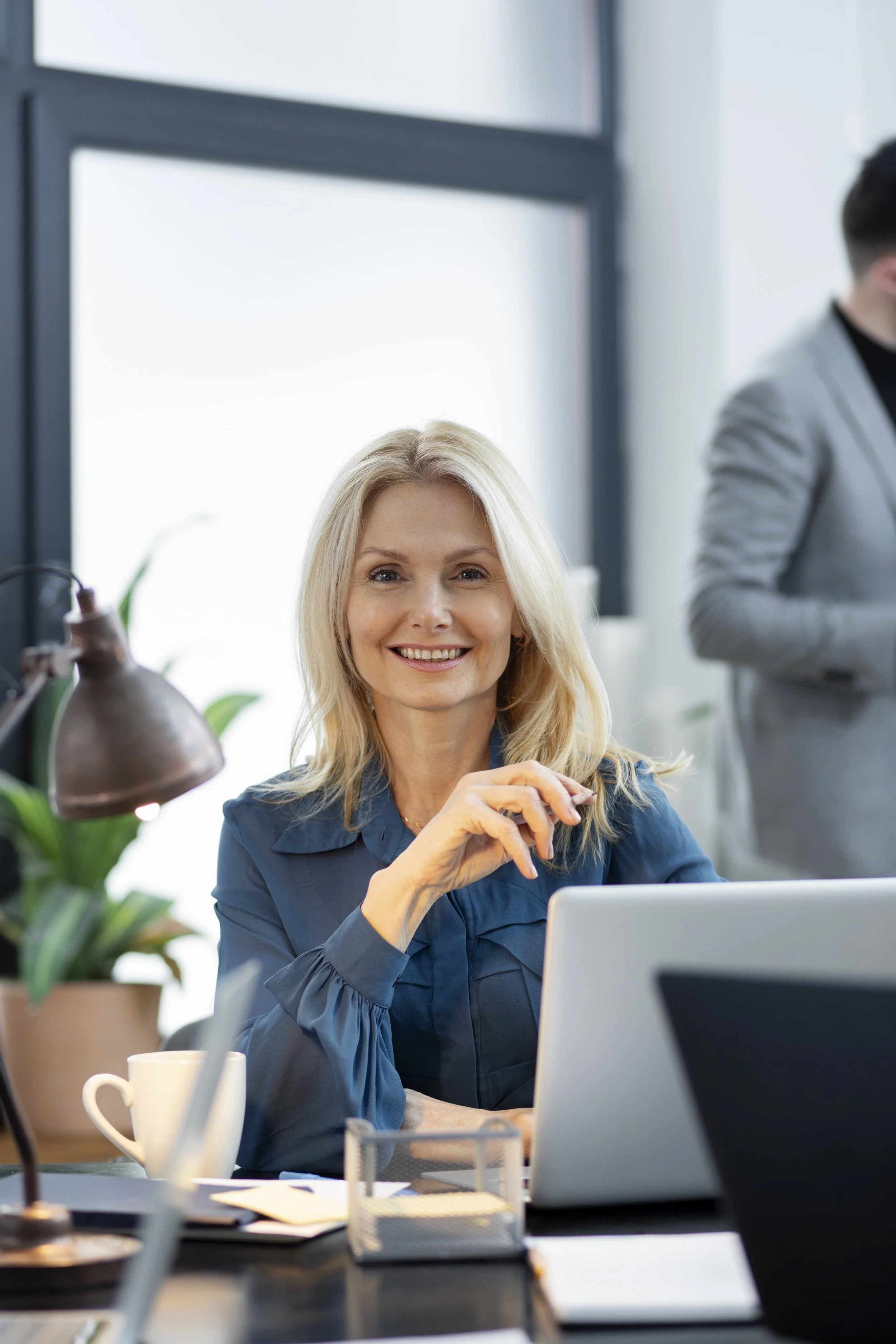 Smiling woman sitting at a desk with a laptop, surrounded by office supplies and plants, in a modern workspace.