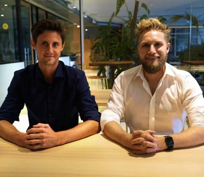 Two men sitting at a table in an office environment, both smiling at the camera, one wearing a dark shirt, and the other in a white shirt with a smartwatch.