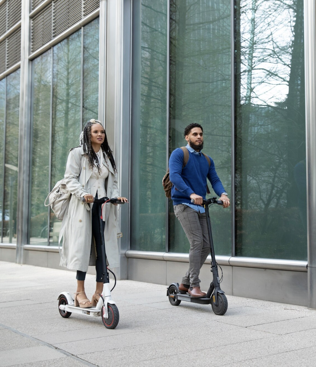 Two people riding electric scooters on a city street with glass buildings in the background.