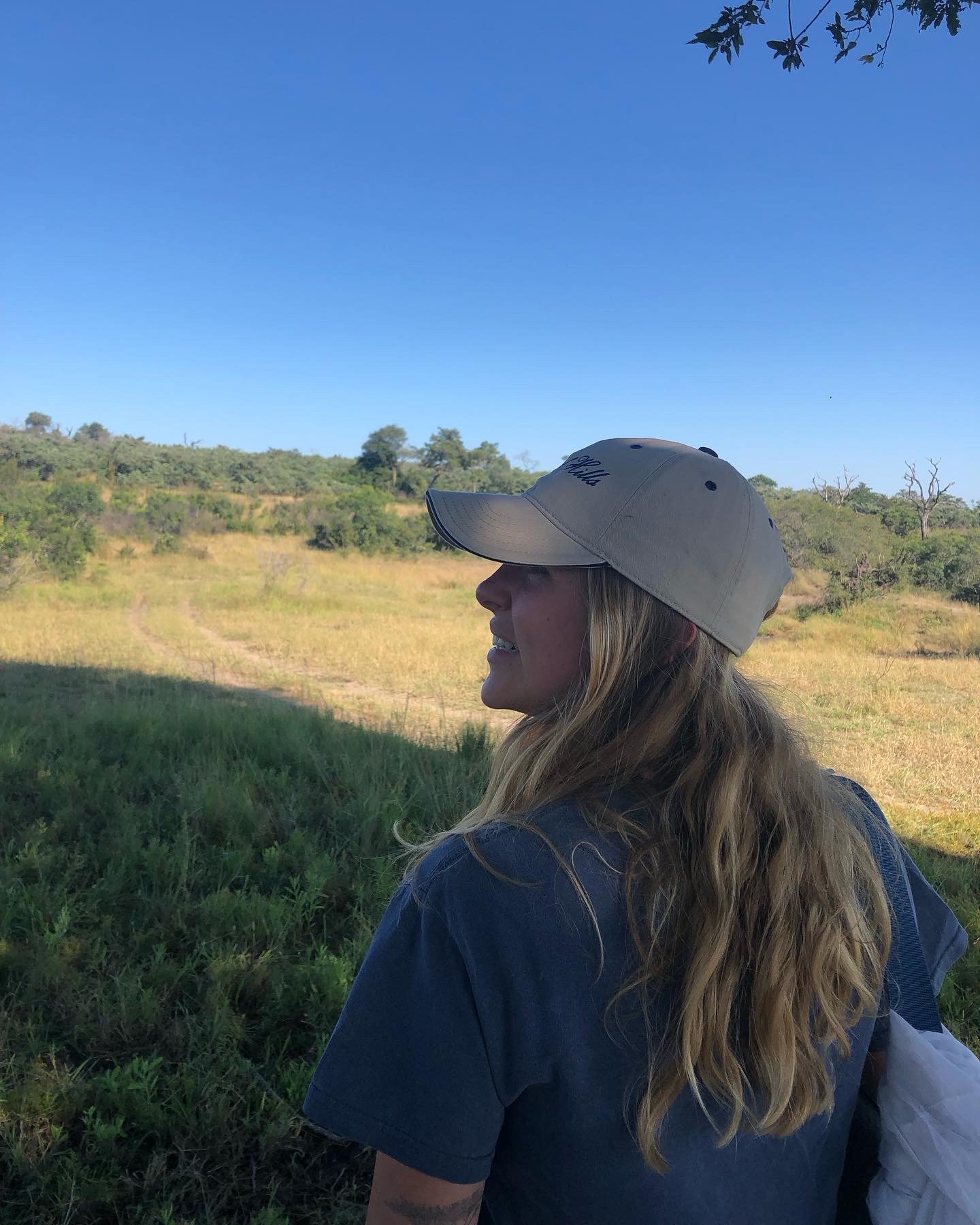 Person wearing a beige cap walking in a grassy field with trees and blue sky.