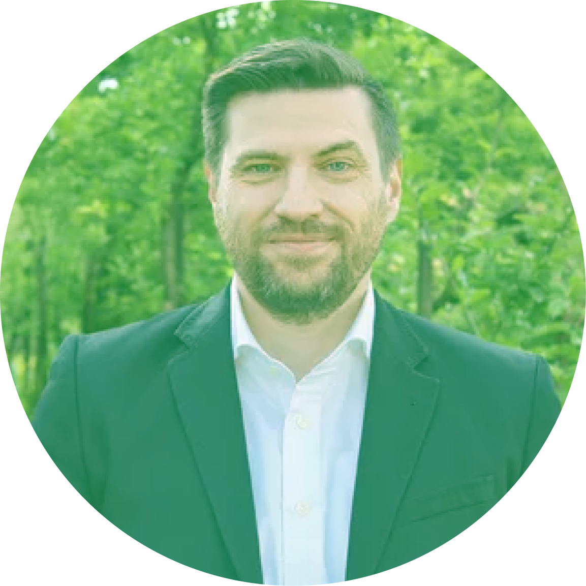 A man in a suit standing outdoors with a background of green trees.