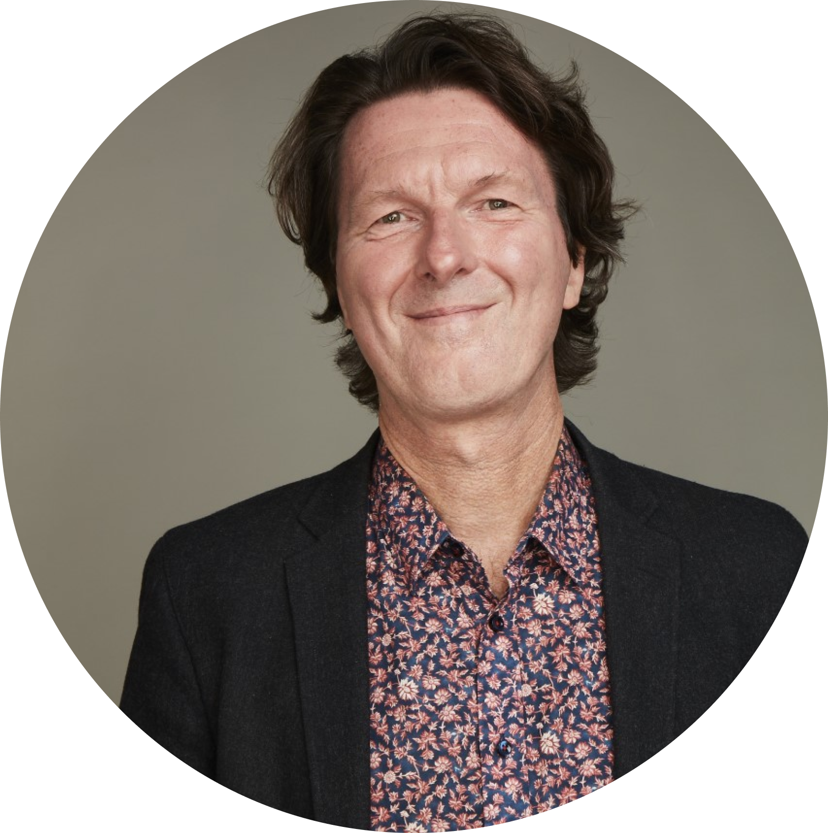 Man with medium-length hair wearing a floral shirt and dark blazer standing against a neutral background, smiling gently.