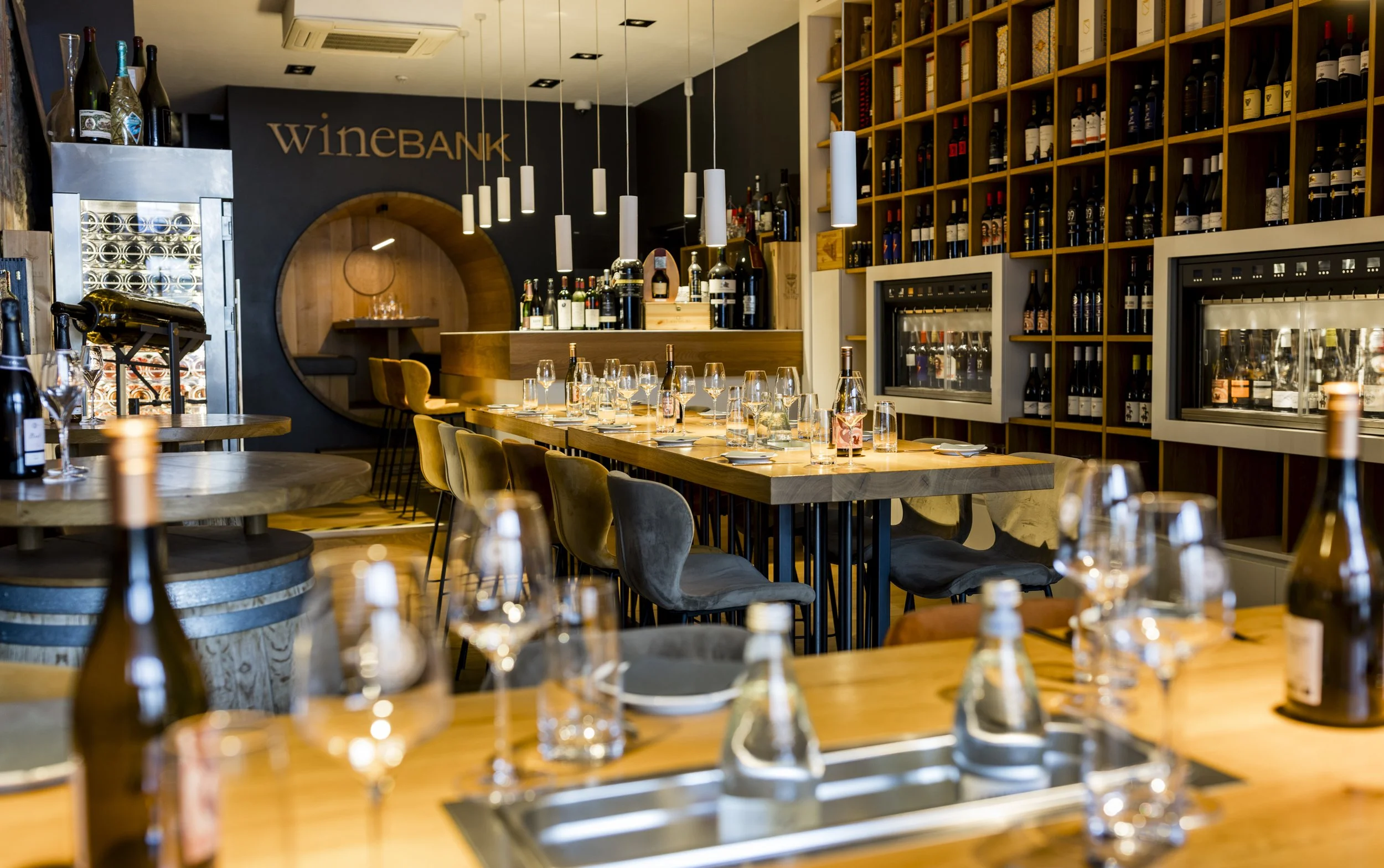 Interior of a modern wine bar with wooden tables, wine bottles, glasses set, and shelves filled with wine. Seating and ambient lighting create a cozy atmosphere.