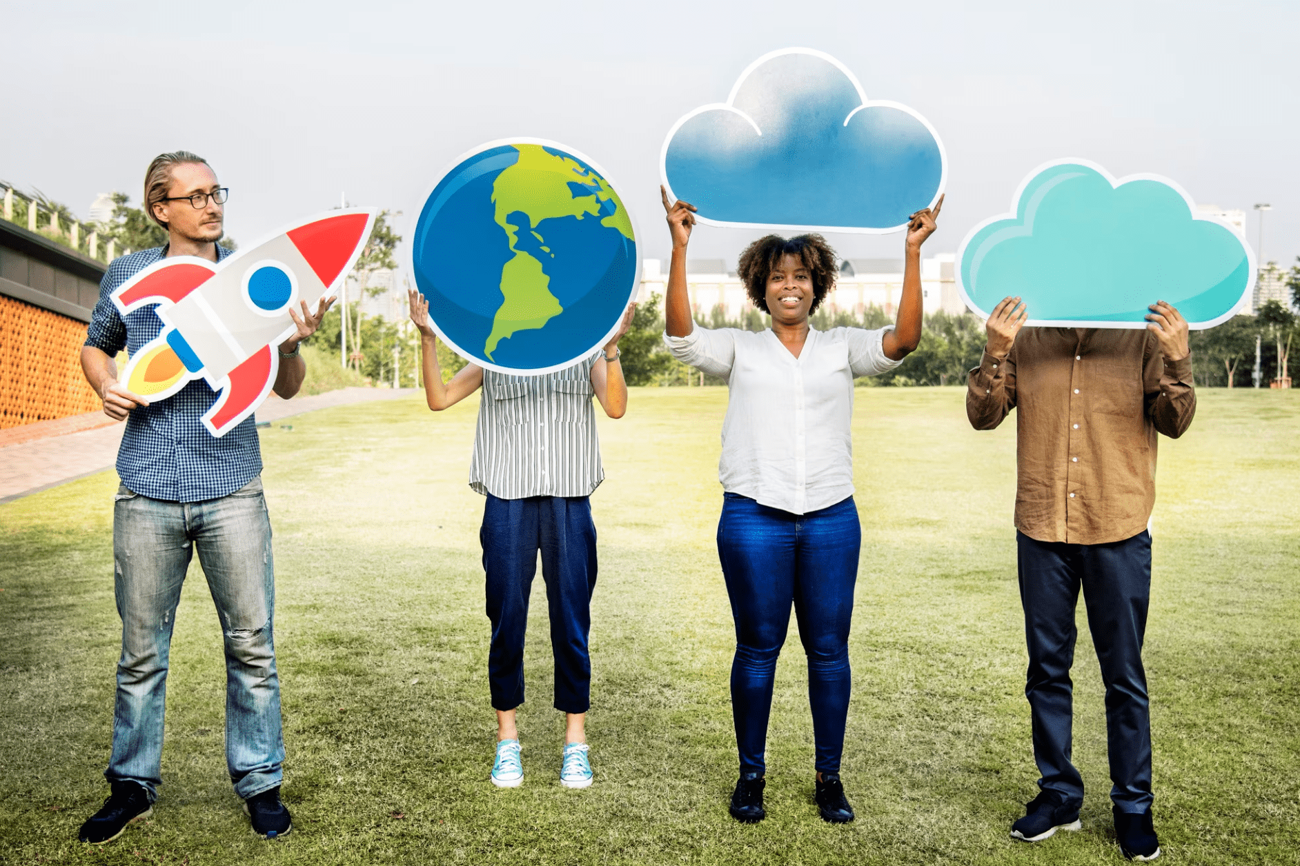 Four people standing in a grassy area holding large cutouts: a rocket, globe, cloud, and another cloud, partially obscuring their faces.