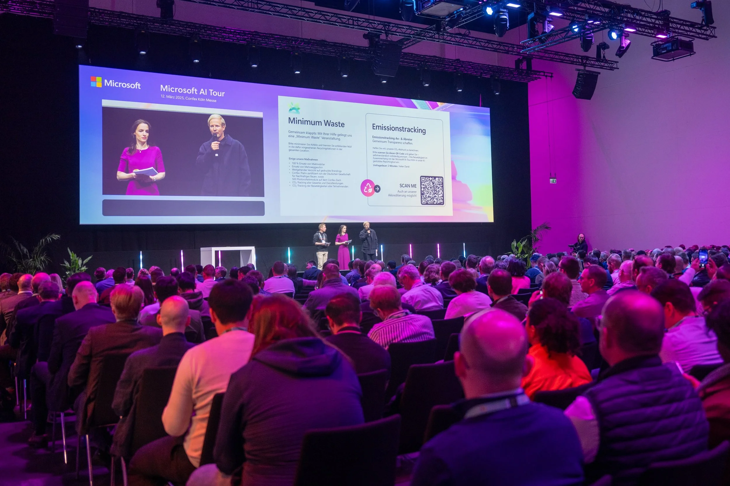 Conference audience watching Microsoft AI Tour presentation, March 12, 2025, in Cologne. Speakers on stage, large screen with presentation on minimum waste and emission tracking. QR code visible on screen.