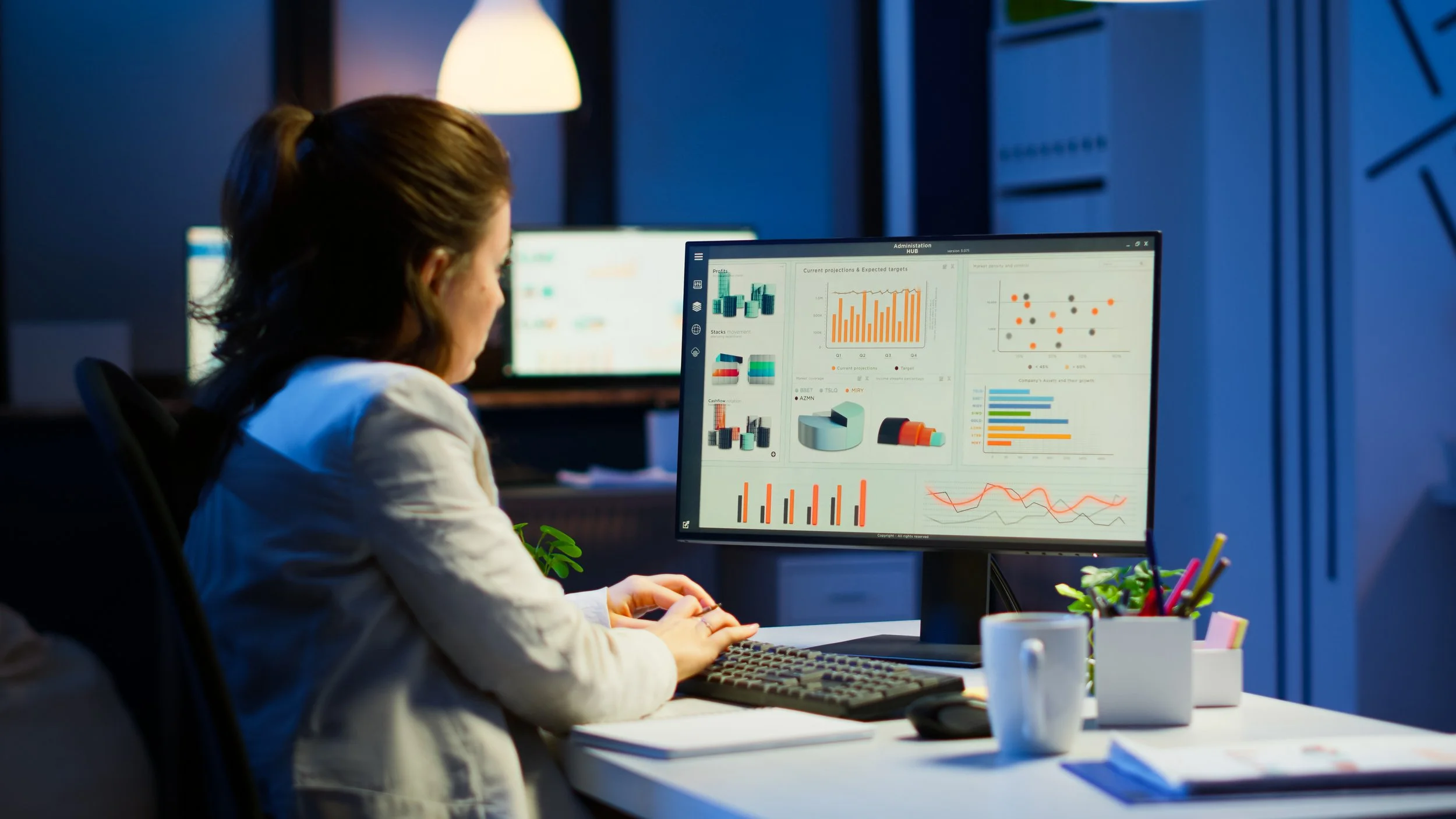 A woman sitting at a desk, analyzing various colorful graphs and charts displayed on a computer monitor in an office setting.