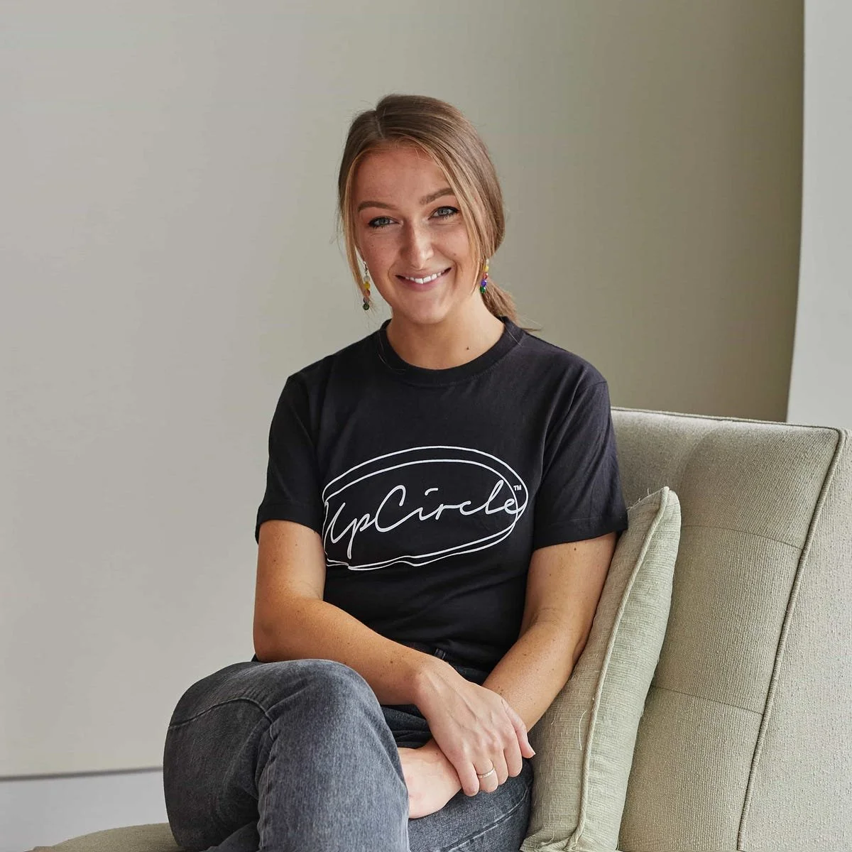 Smiling person sitting on a chair, wearing a black "UpCircle" t-shirt and jeans, with hair tied back and colorful earrings.