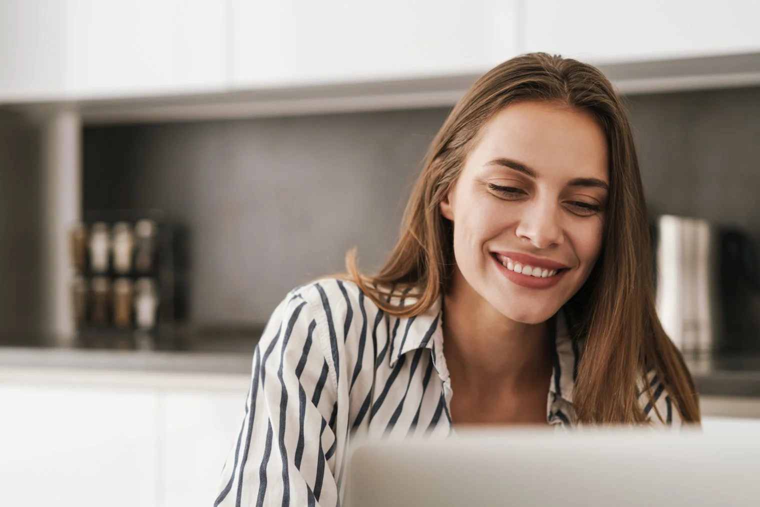 Woman smiling in a kitchen, looking at a laptop.