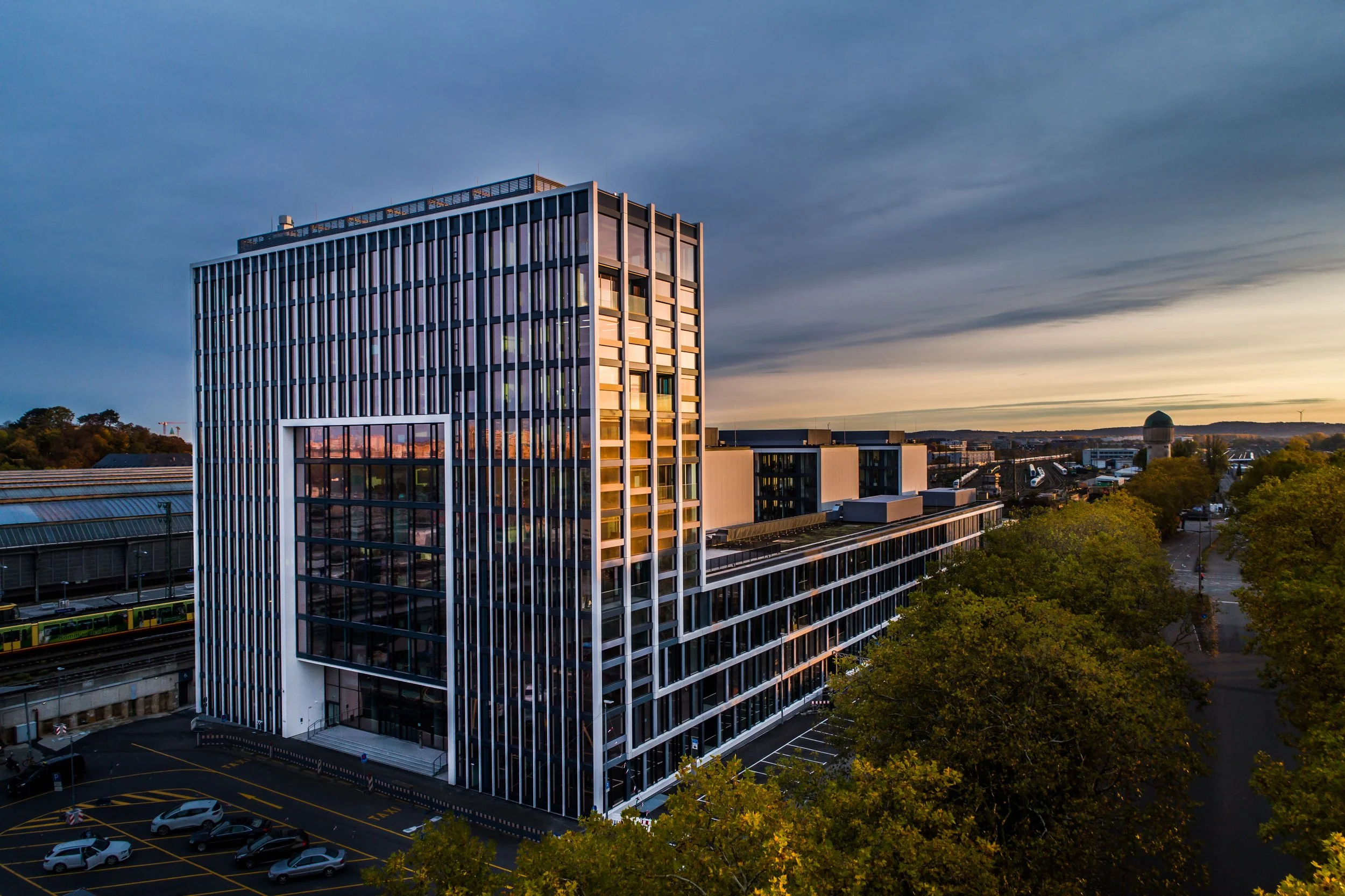 Modern office building with glass facade at sunset, surrounded by trees and parking lot; city skyline in background.