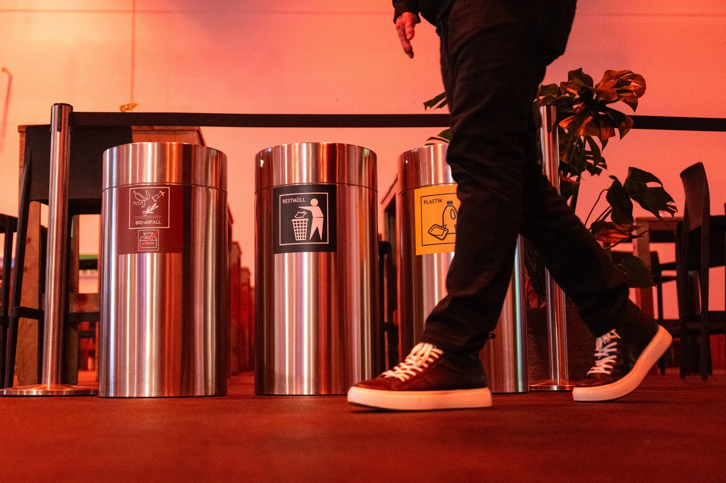 Stainless steel recycling bins labeled for sorting waste into organic, residual, and plastic categories, with a person walking by.