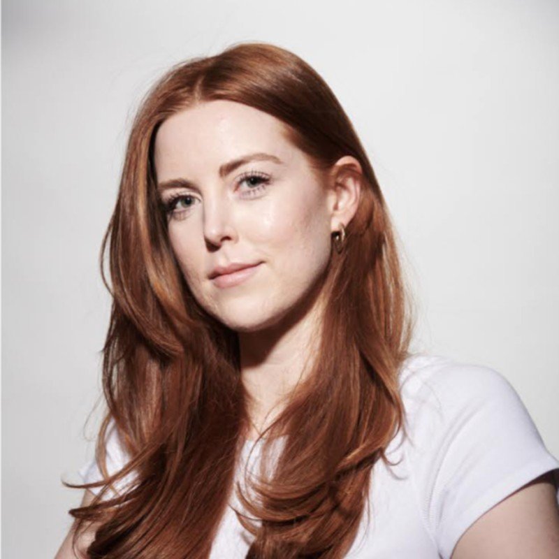 Woman with long red hair wearing a white top, looking towards the camera on a light background.
