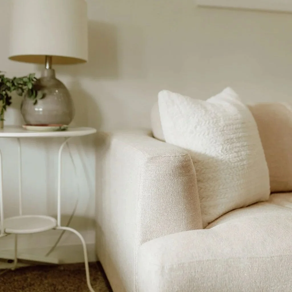 Cozy cream sofa with textured white pillows beside a round white side table, lamp, and potted plant