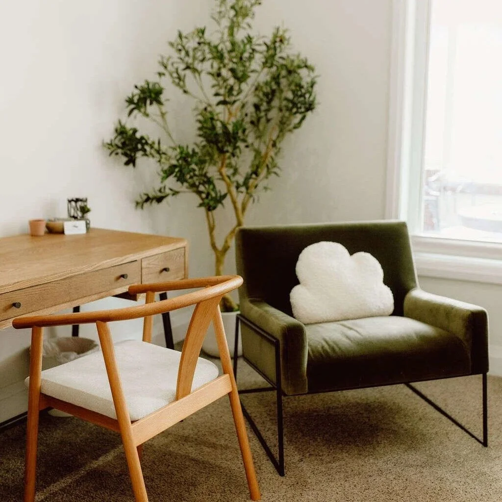 Cozy modern living room with a wooden armchair, olive green lounge chair, cloud-shaped pillow, dresser, and tall indoor tree by a window