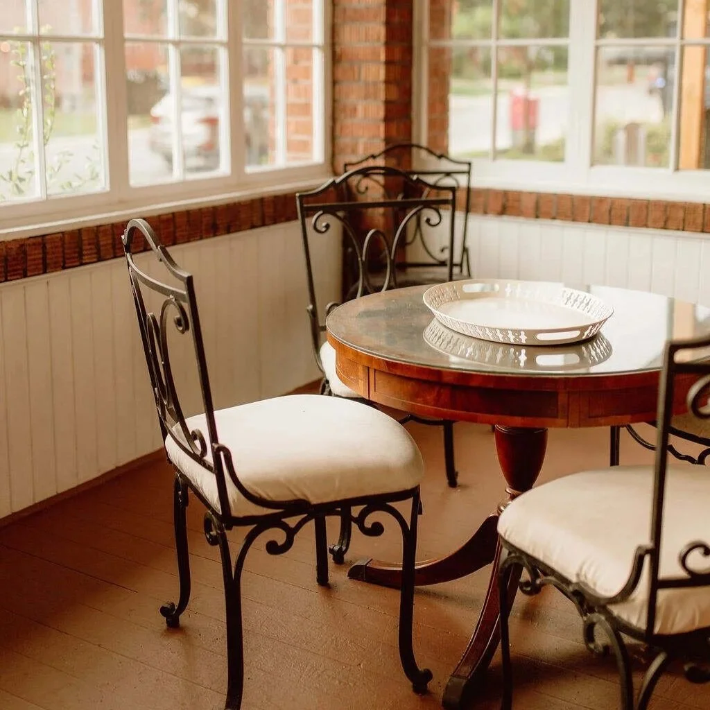 Sunlit dining nook with a round wooden table, white bowl, and ornate wrought-iron chairs by large windows.