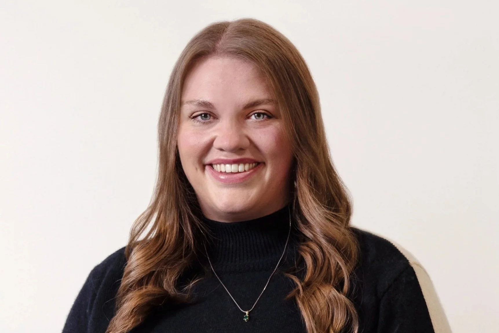 Smiling person with brown hair wearing a black sweater and necklace, against a plain white background.