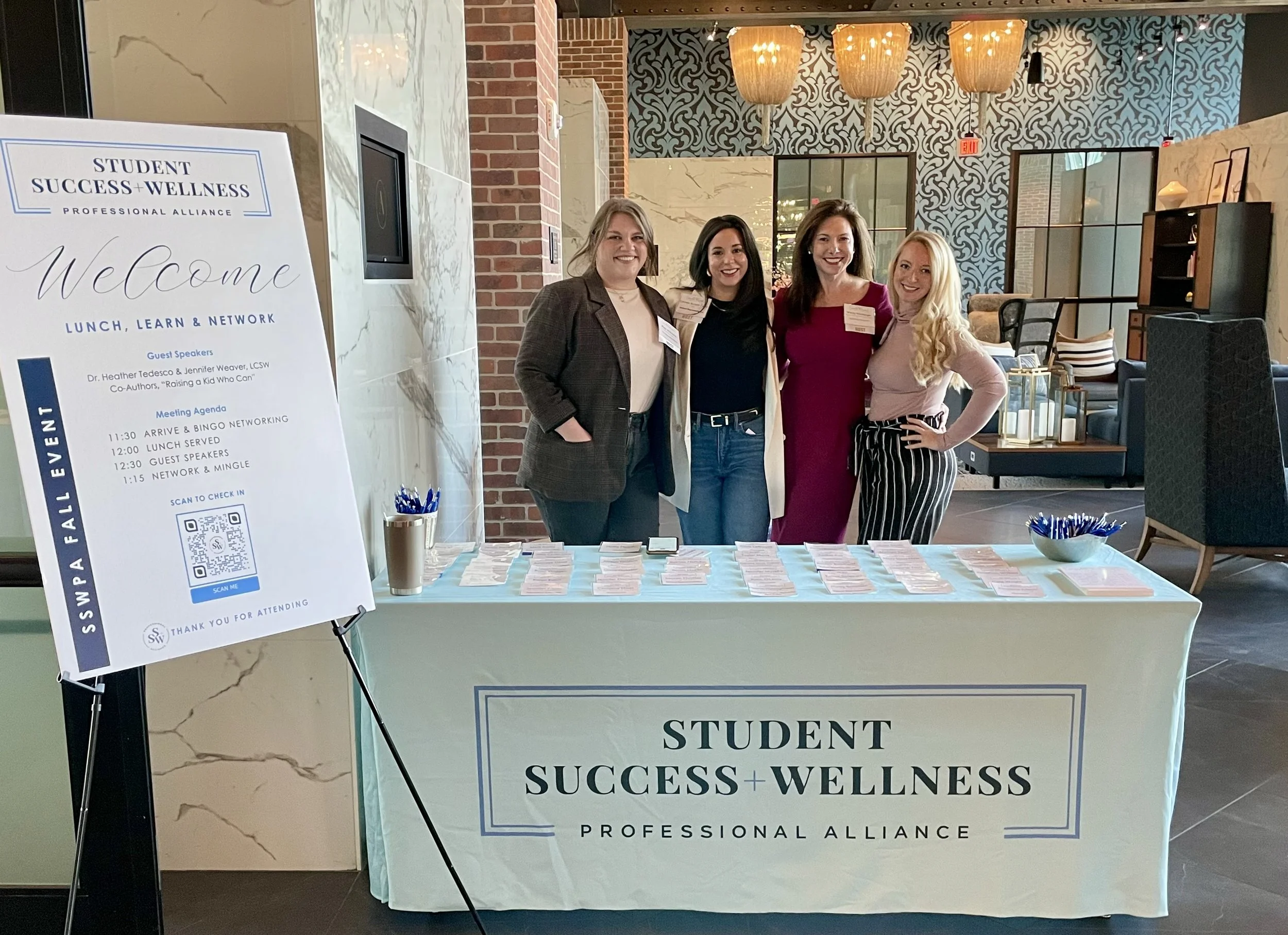 Four women standing behind a table at a wellness event, smiling, with a large sign promoting 'Student Success + Wellness' in front of them. The table has name tags and pens, and the background features a modern interior with decorative lighting and furniture.