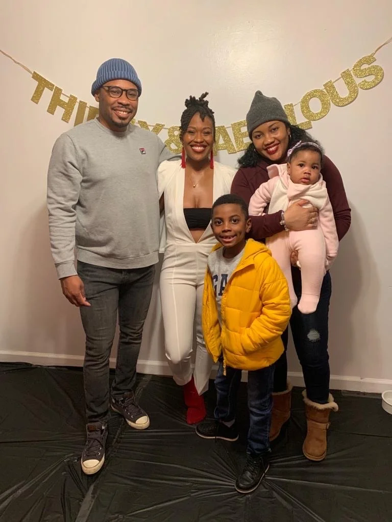 Group of five people smiling in front of a "Thirty & Fabulous" banner, including two adults, a teen, a young boy in a yellow jacket, and a baby in pink.