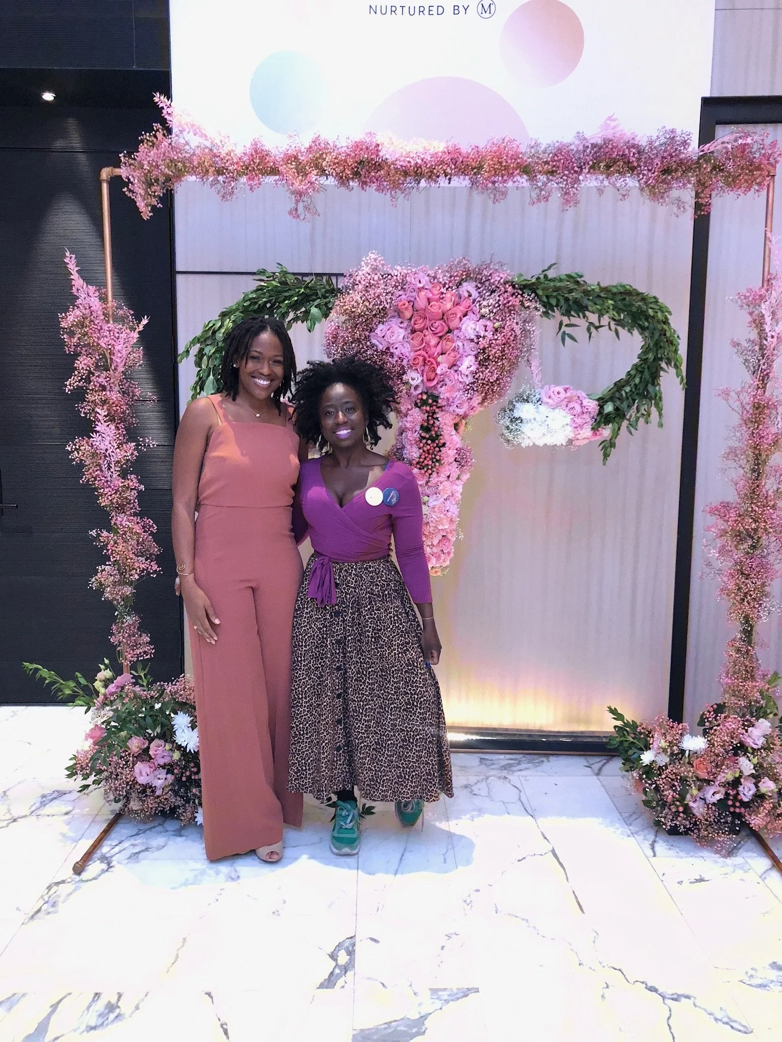 Two people pose in front of a decorative floral display. The display features pink and green flowers arranged in the shape of a uterus. The person on the left wears a sleeveless rust-colored jumpsuit, while the person on the right wears a purple top,