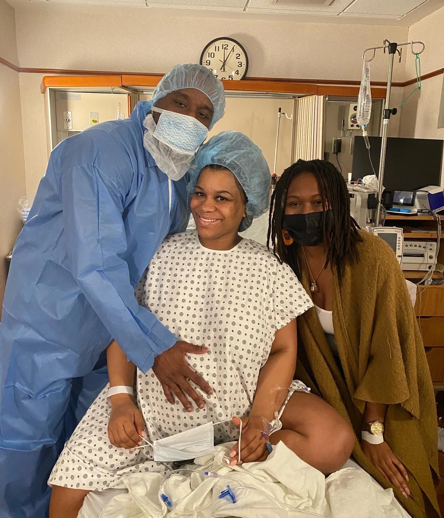 A woman in a hospital gown sitting on a hospital bed, smiling, with a man and another woman standing beside her. The man is wearing blue scrubs and a surgical mask, and the other woman is wearing a brown sweater and face mask. Medical equipment is vi