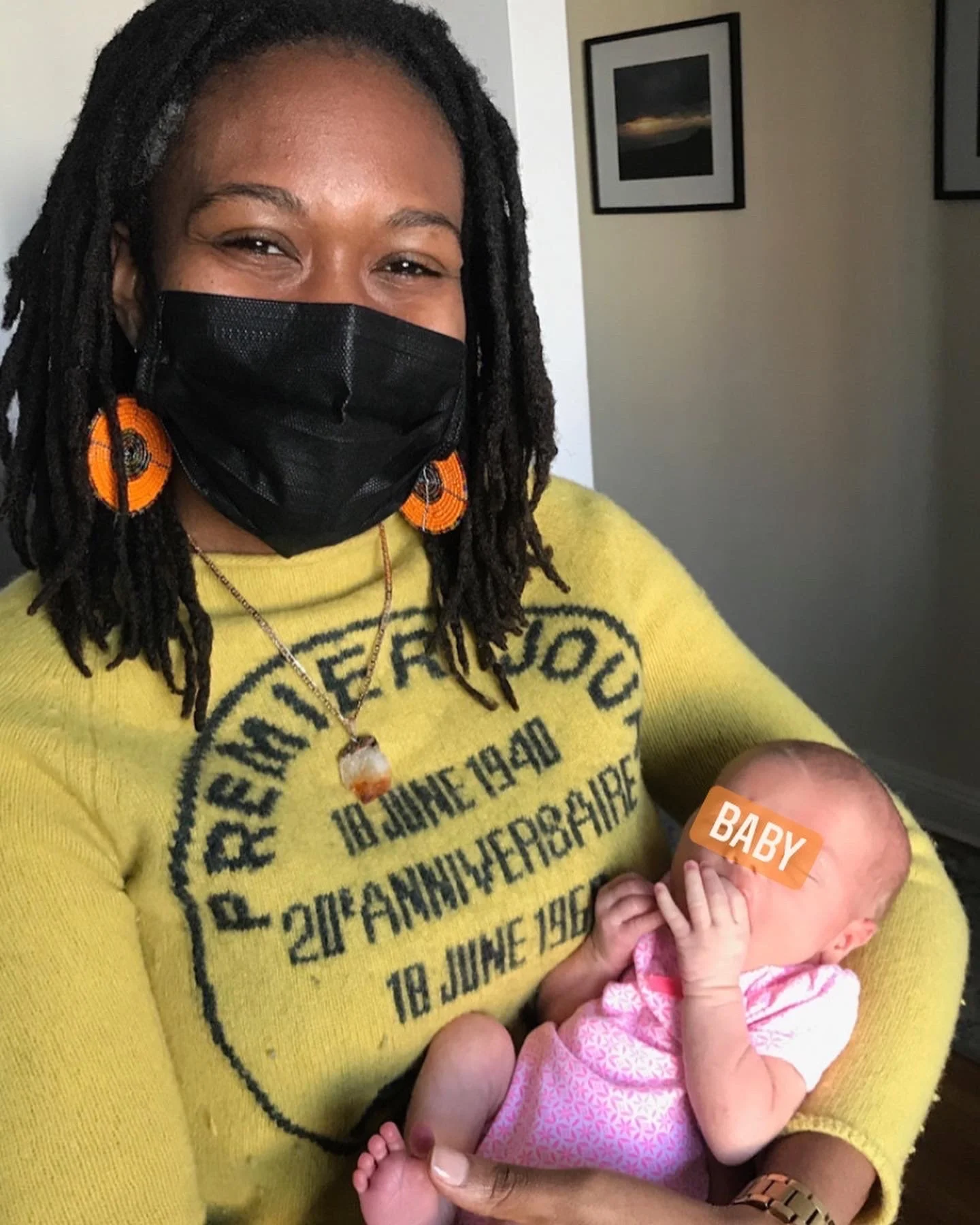 Masked person holding a newborn baby with "BABY" sticker on face, wearing a yellow sweater and orange earrings.