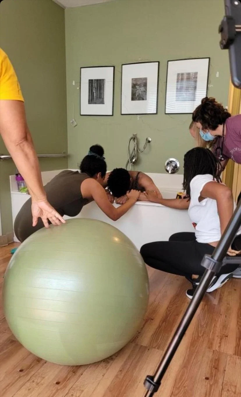Women supporting a person in a bathtub during labor, with an exercise ball nearby in a birth center setting.