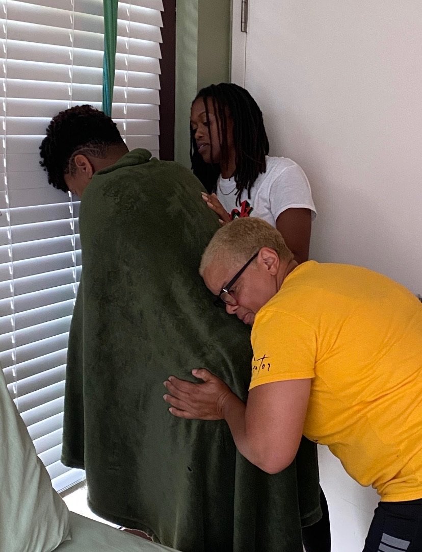 Three people hugging near a window, one wrapped in a green blanket.