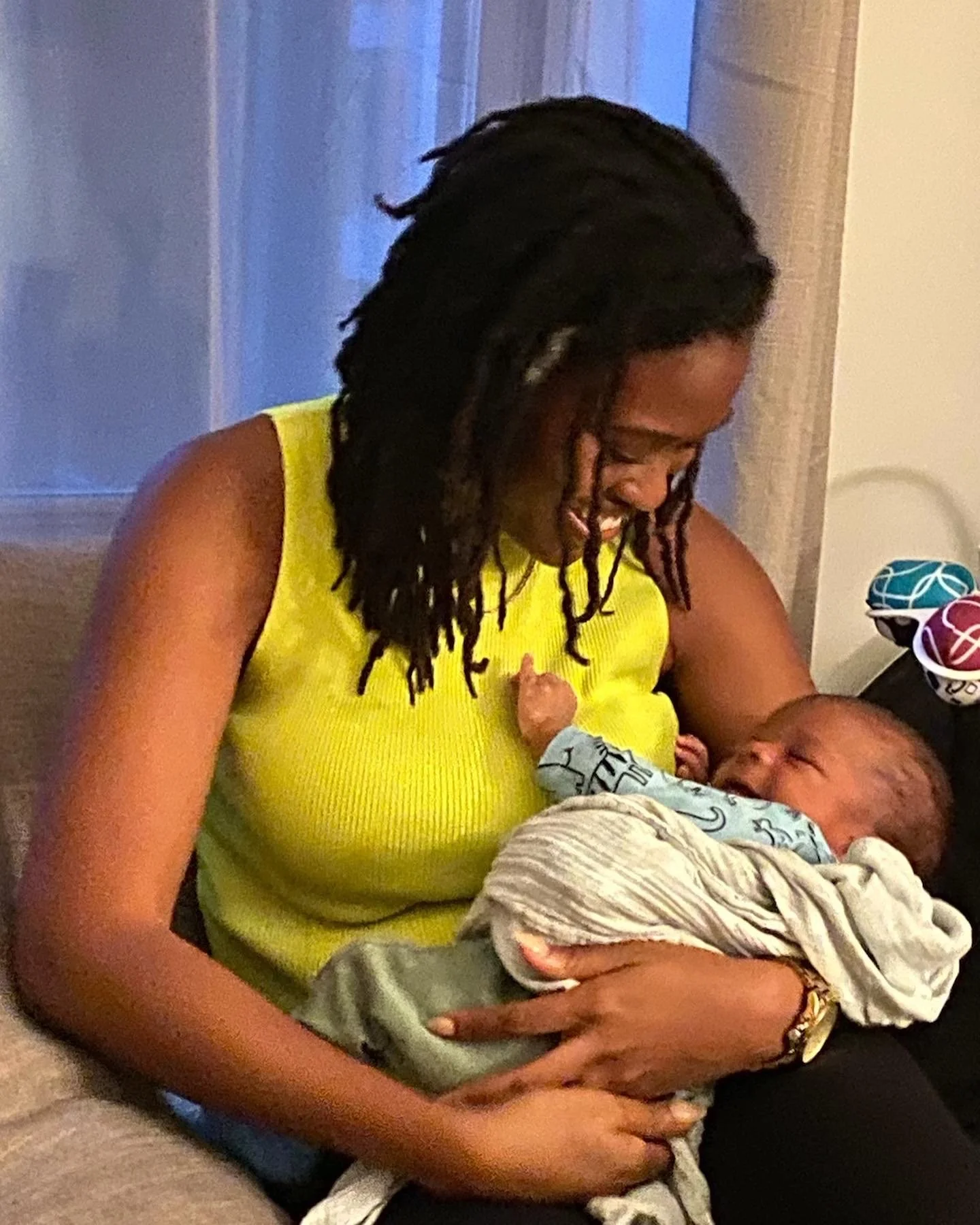 A woman with long hair wearing a sleeveless yellow top holding a sleeping baby wrapped in a blanket.