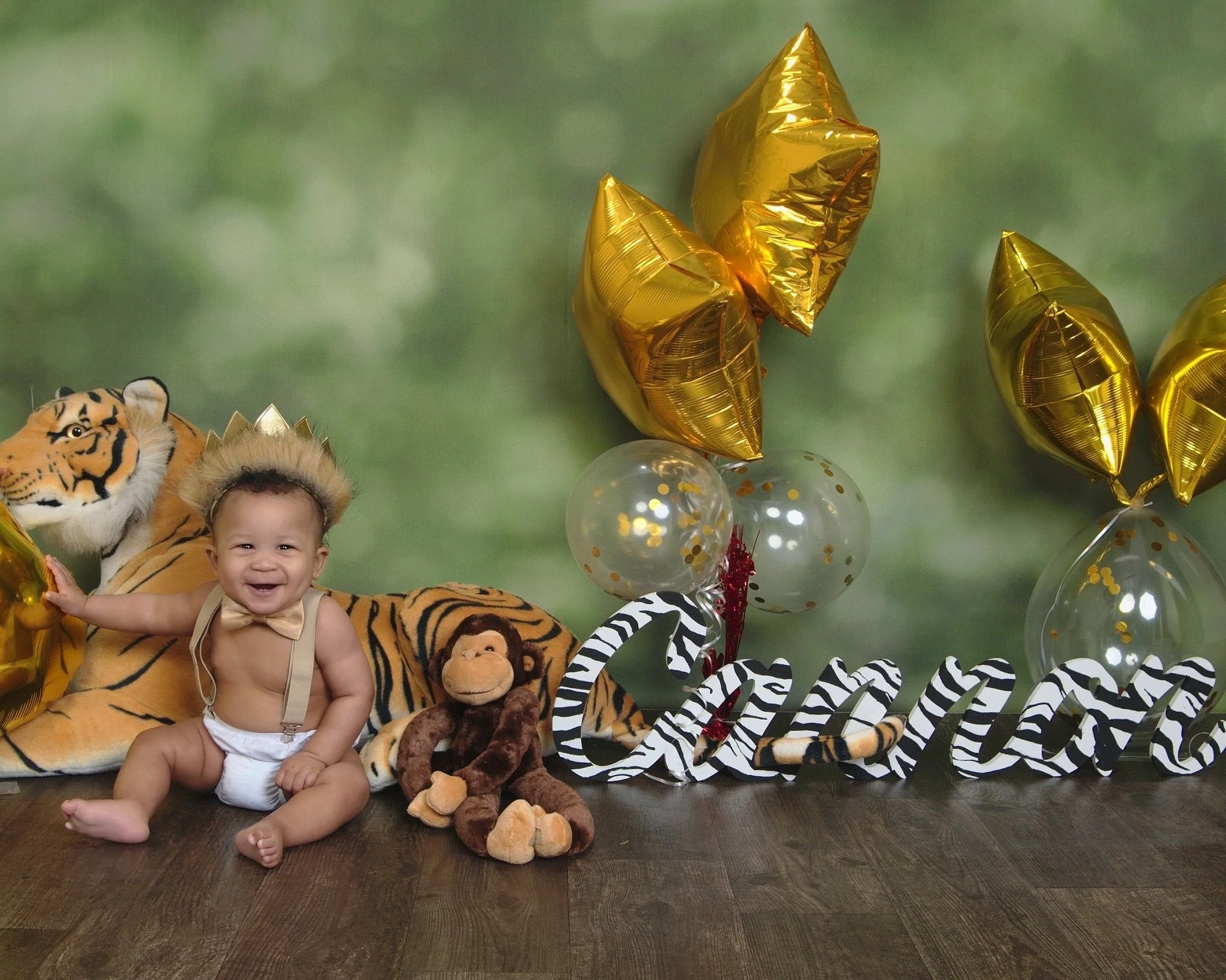A baby wearing a gold crown and bow tie sits on the floor, surrounded by plush animal toys, including a large tiger and a monkey. Nearby, gold star balloons and zebra-striped letters spell out a name. The background is a green, leafy pattern.