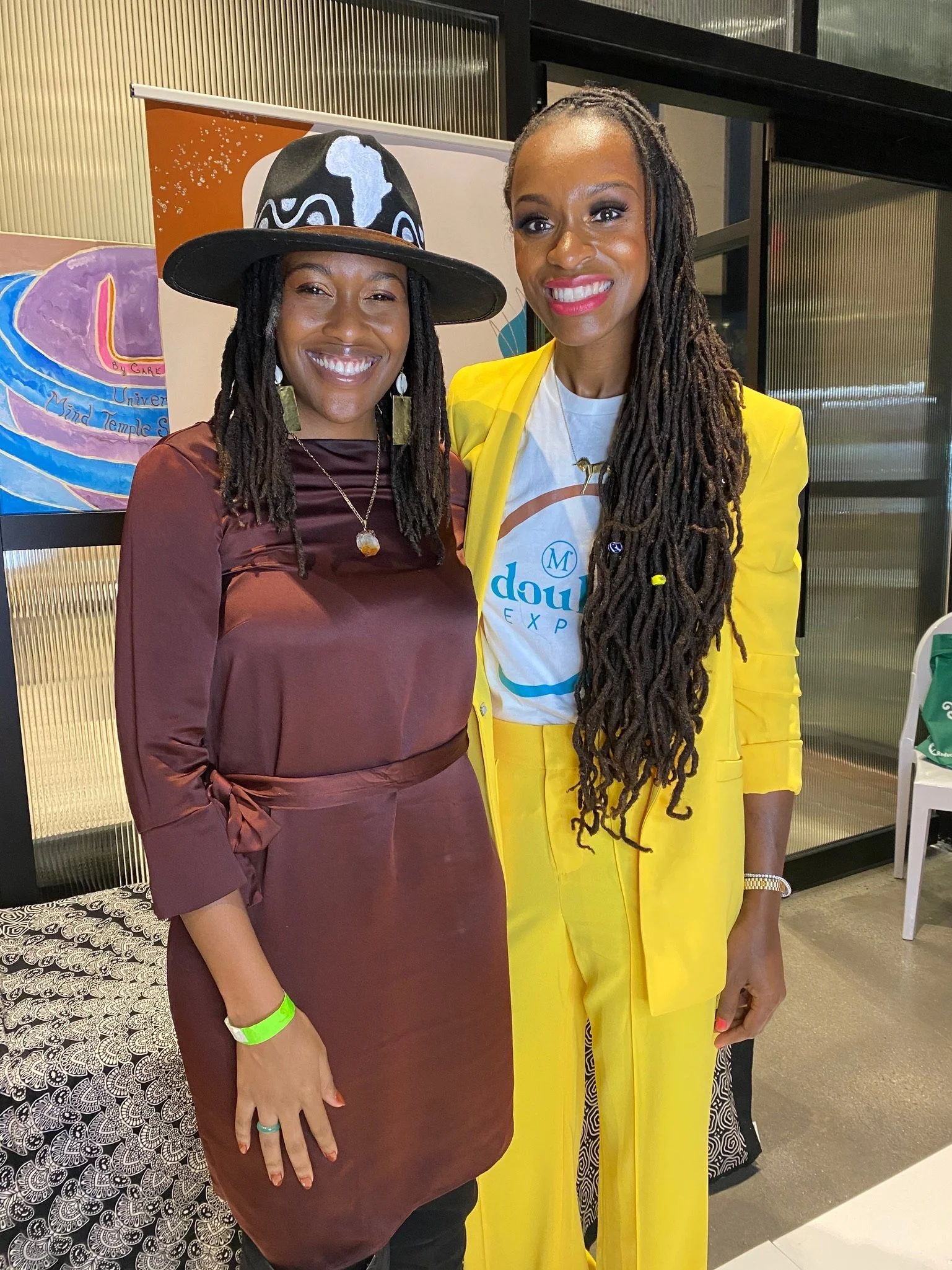 Two women smiling at an event; one wearing a black hat with white designs and a brown dress, the other in a bright yellow pantsuit.