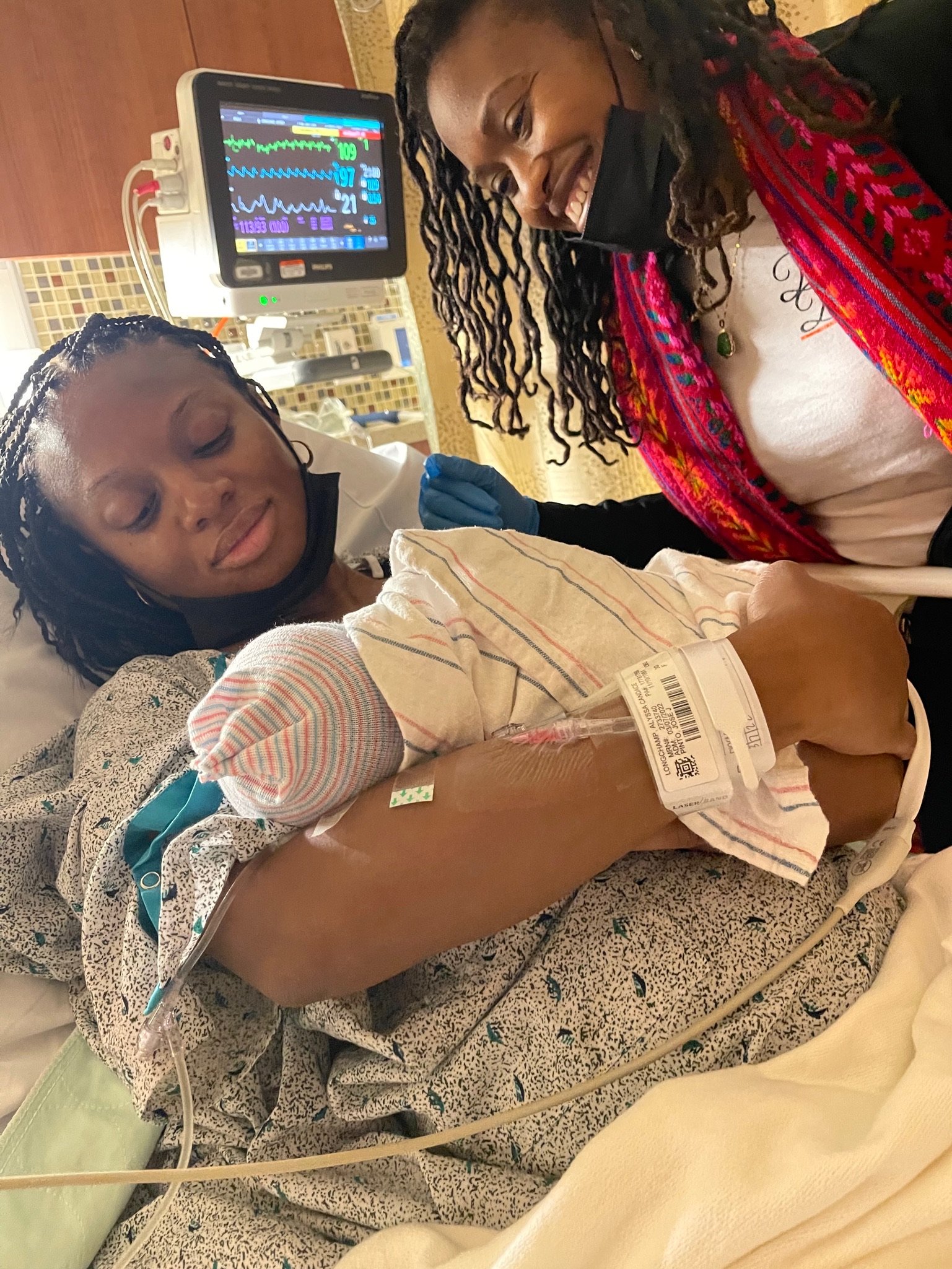 New mother holding newborn baby in hospital bed with a friend smiling nearby, medical monitor in the background.